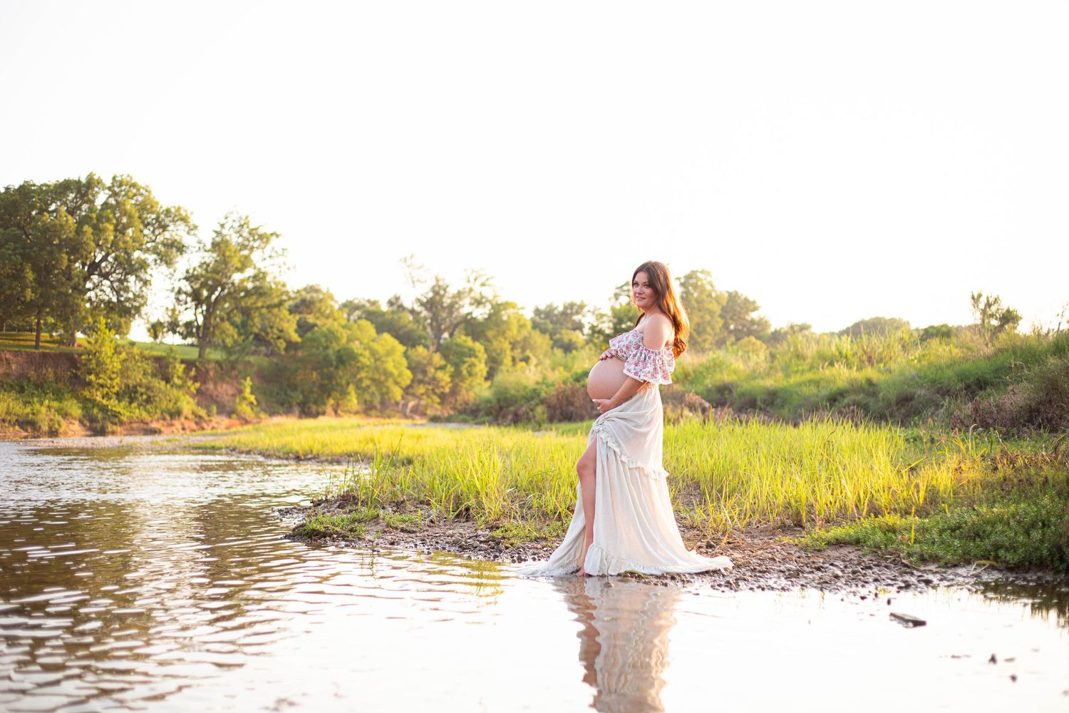 Pregnant woman in white gown standing in shallow water at golden hour with lush greenery background.