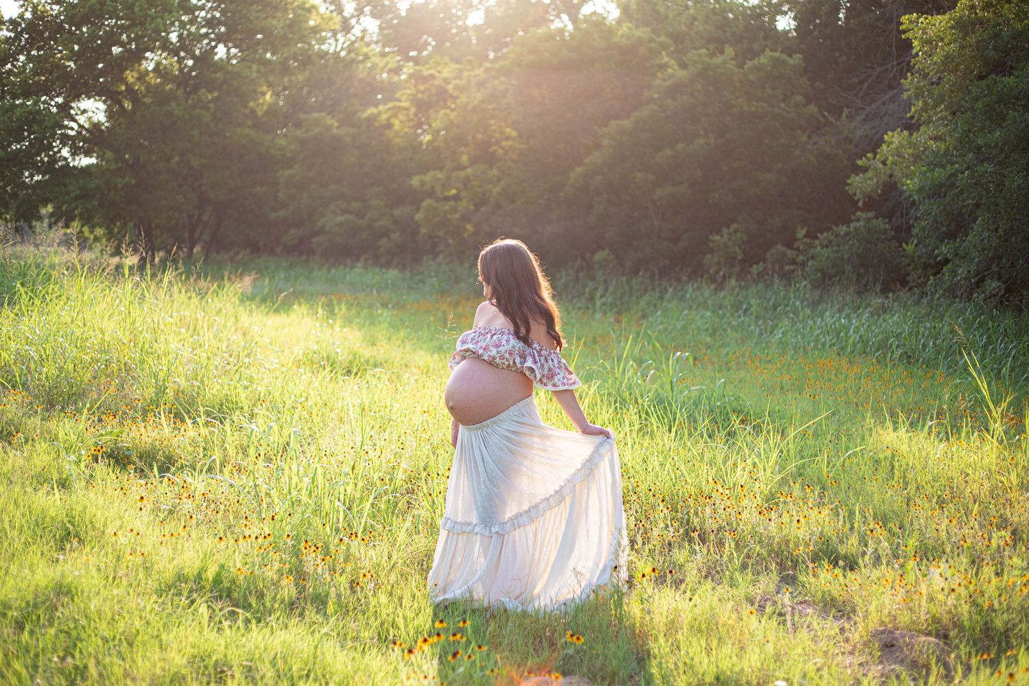 Pregnant woman in white dress standing in sunlit meadow surrounded by lush green trees during golden hour.