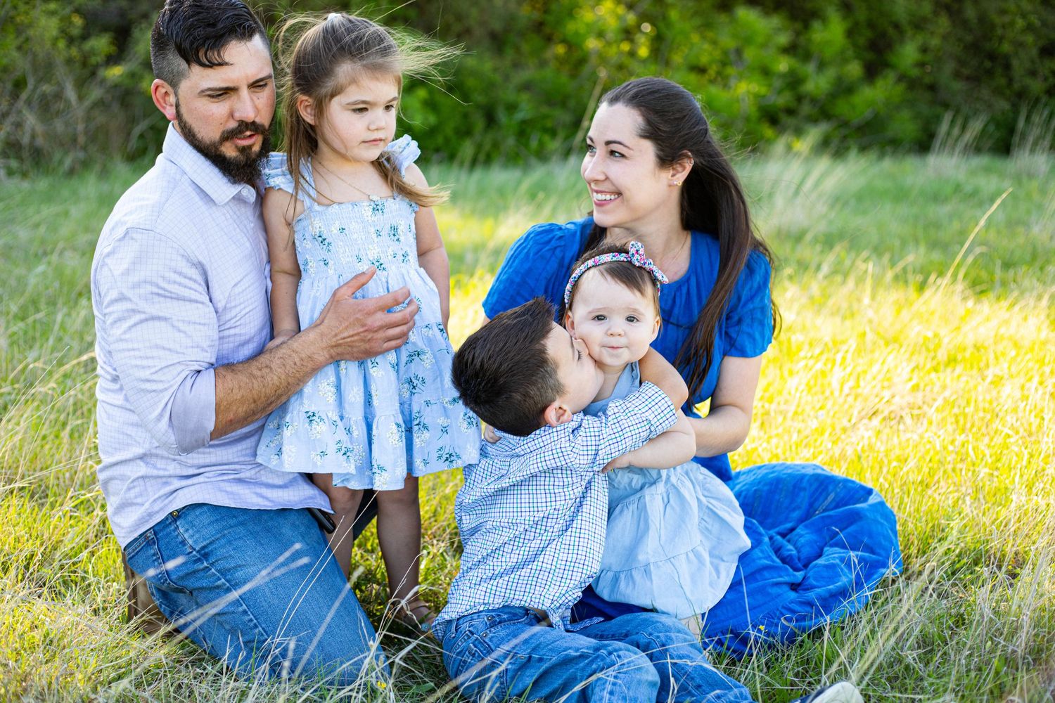Family of four sitting in a green meadow, parents and two young children in coordinating blue outfits during outdoor photo session.