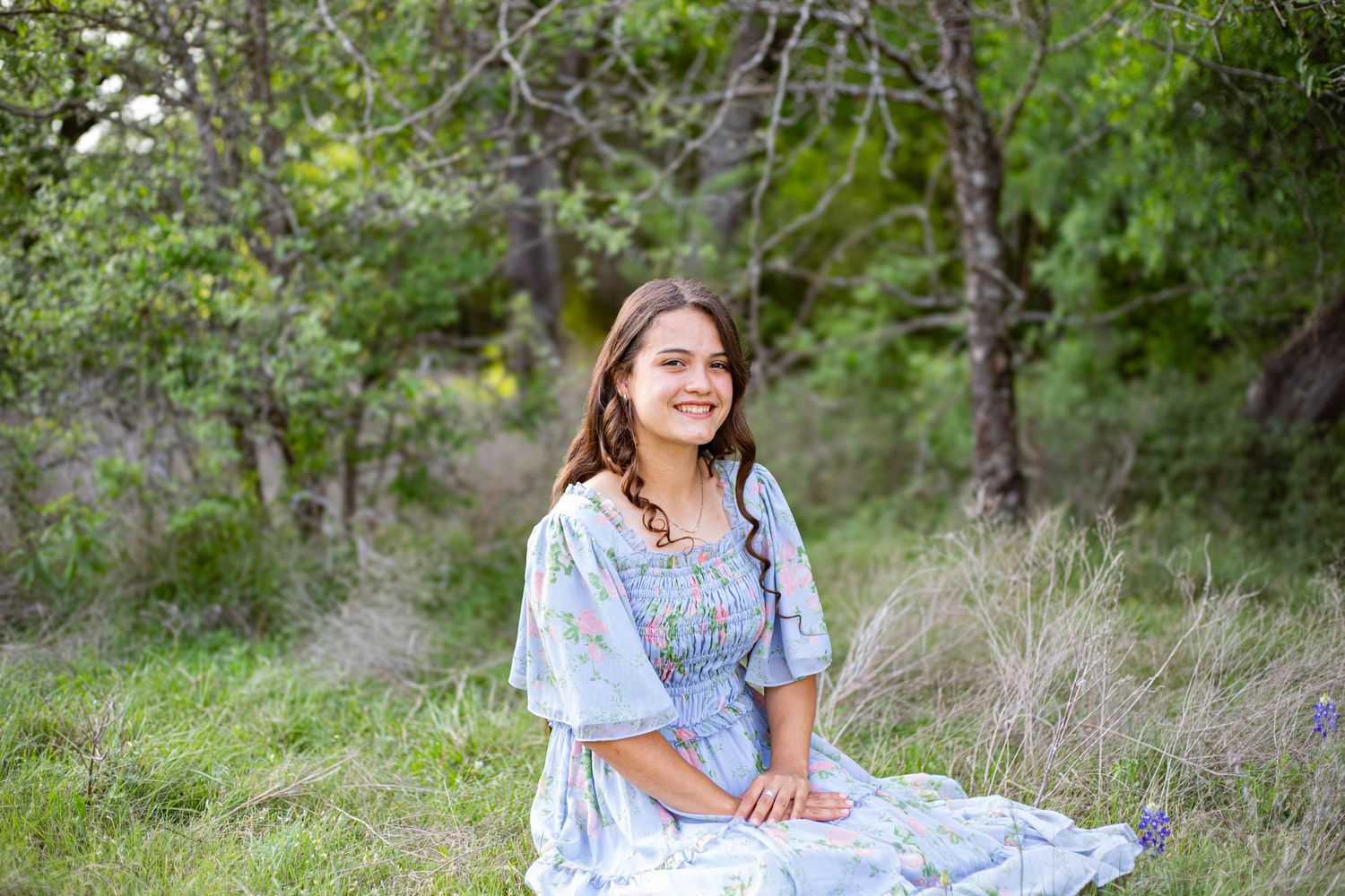 Young woman in a floral blue dress sitting in a lush green meadow with trees, smiling outdoors.