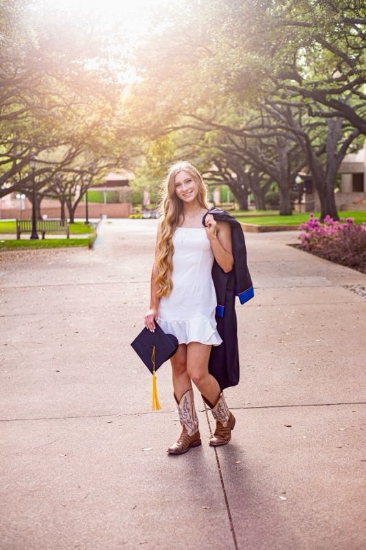 Graduate in cap and gown poses playfully on a tree-lined path during golden hour.