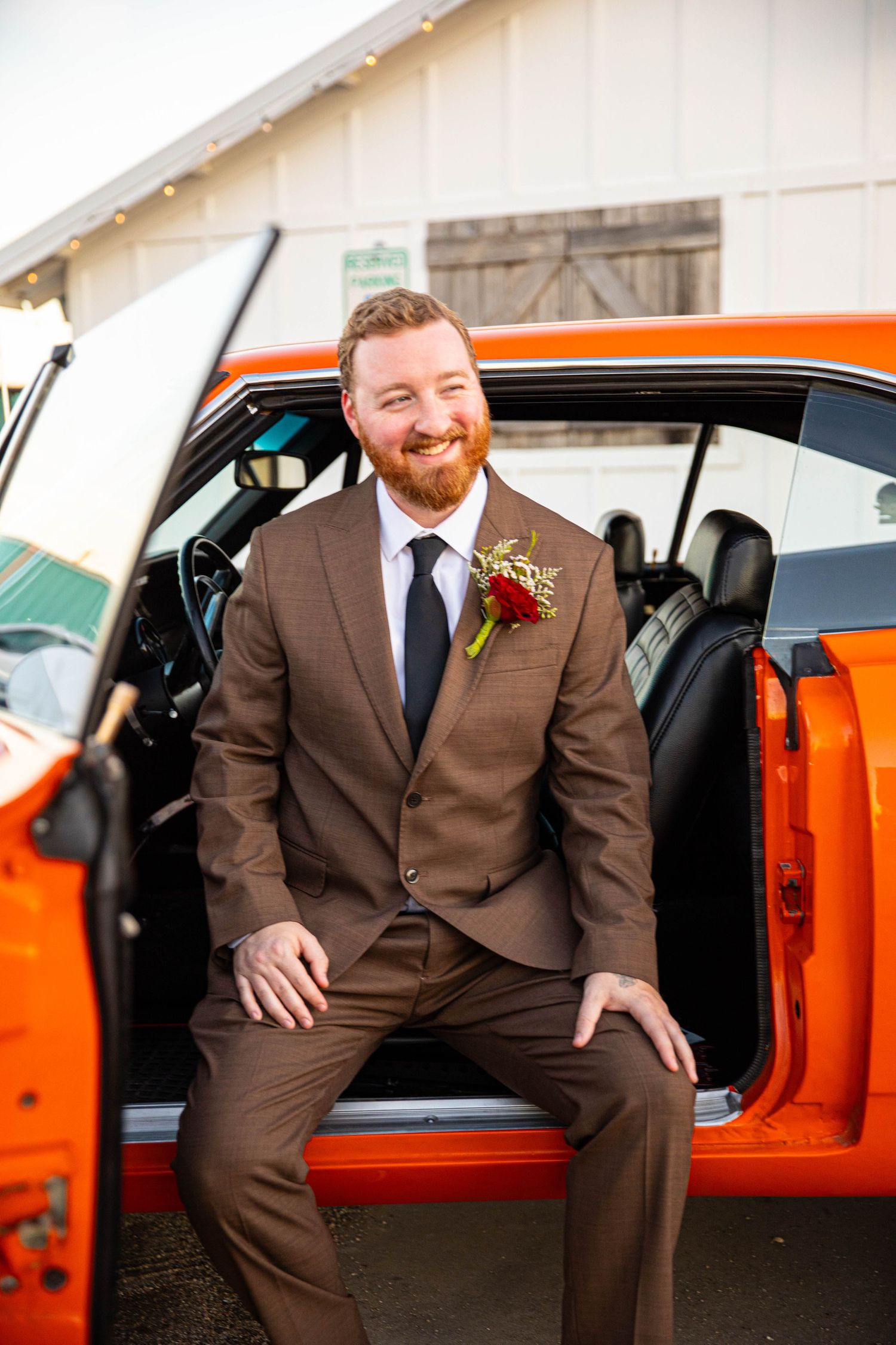Smiling groom in brown suit with boutonniere sits in orange classic car doorway at rustic barn wedding venue.