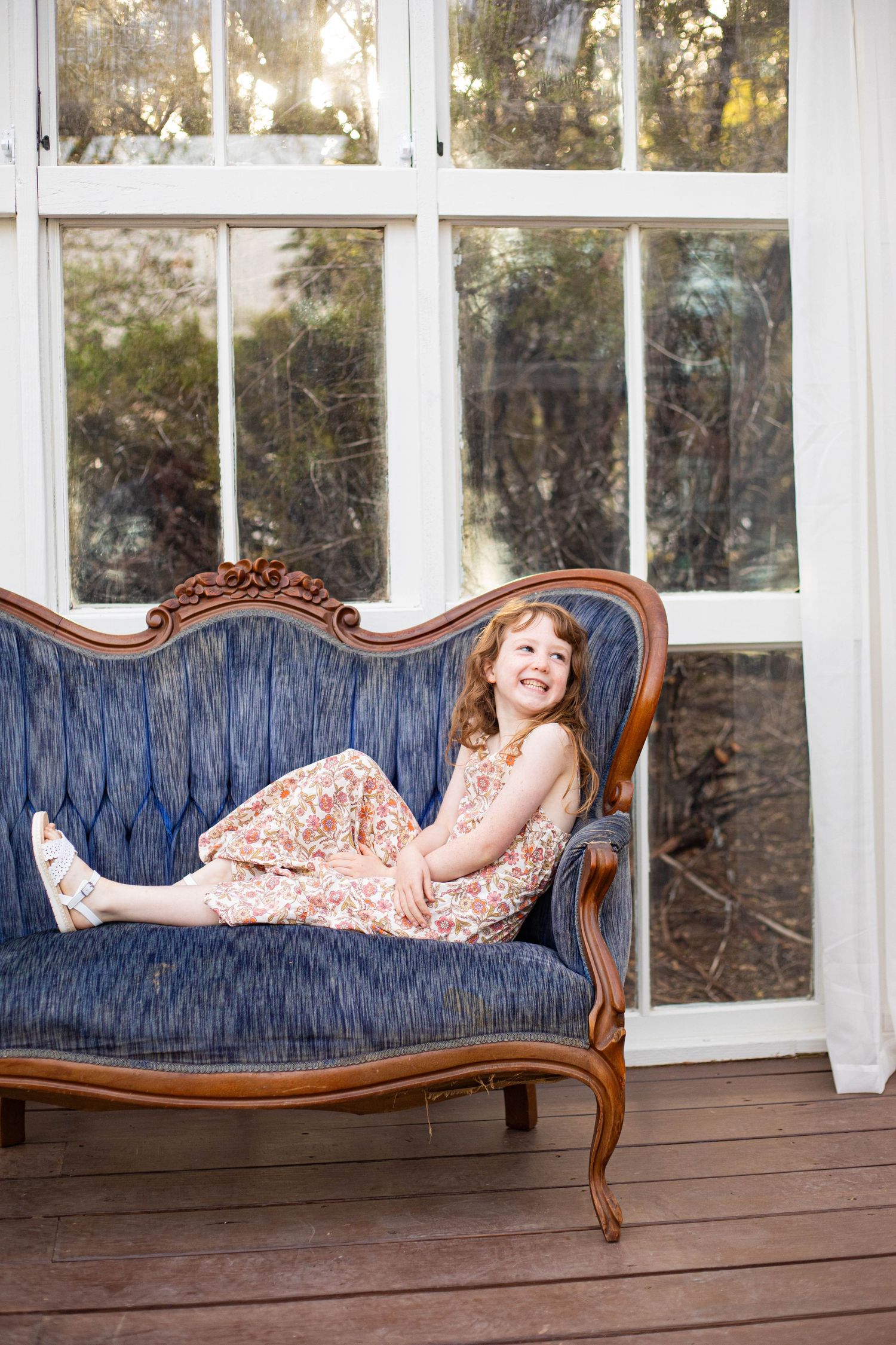A cheerful young girl in a floral dress relaxing on a vintage blue velvet sofa near large windows.