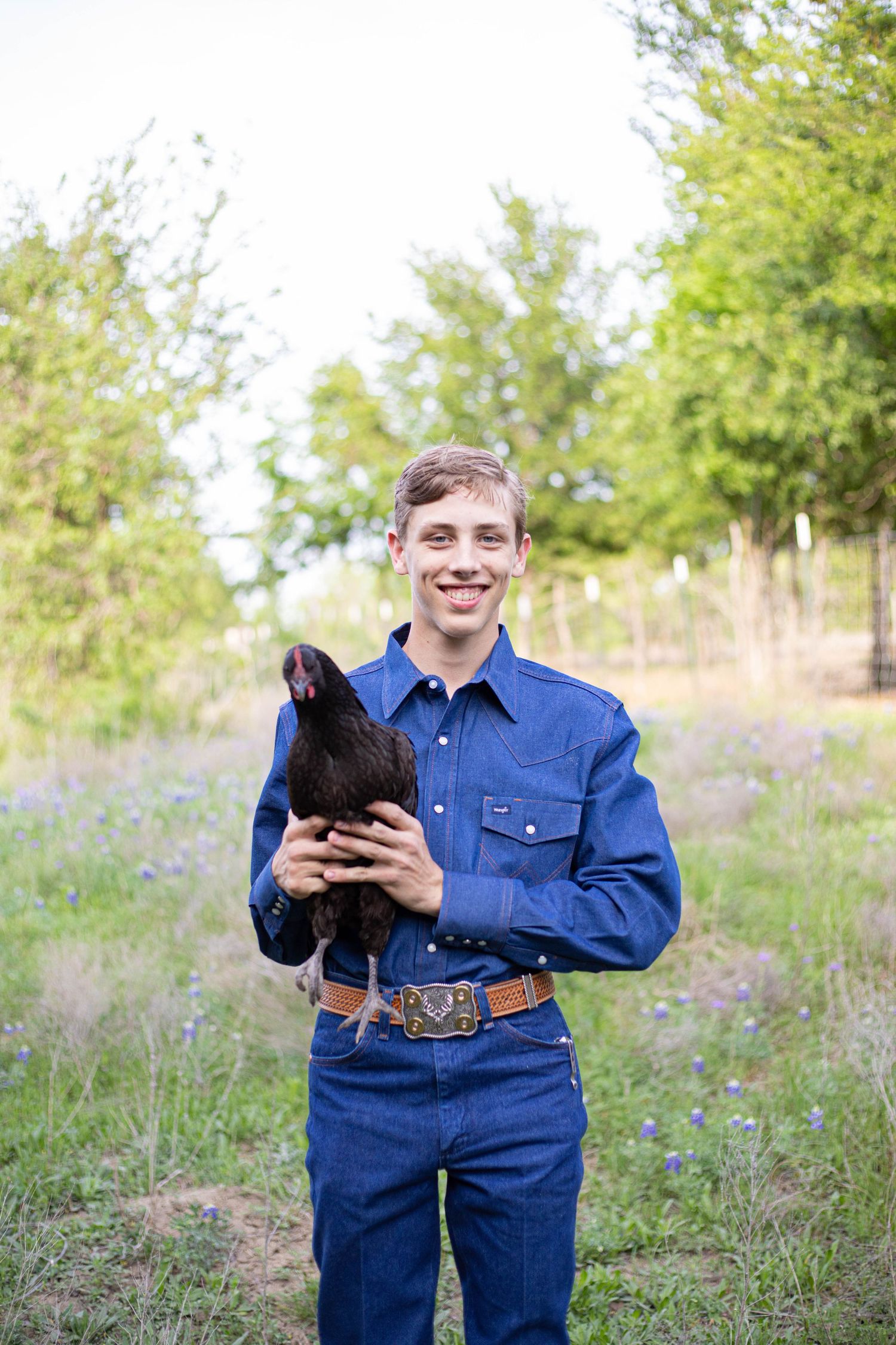 Young man in denim outfit smiling while holding a black chicken outdoors among bluebonnets and green trees.
