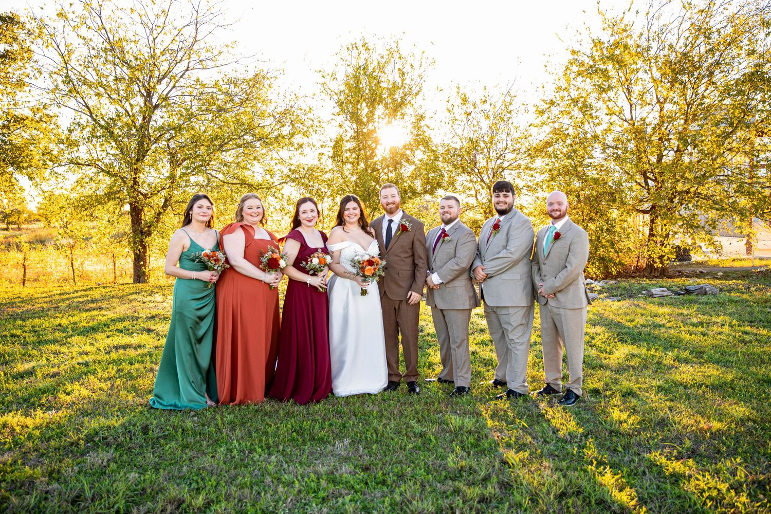 A wedding party in burgundy dresses and grey suits poses together on a grassy hill at sunset.