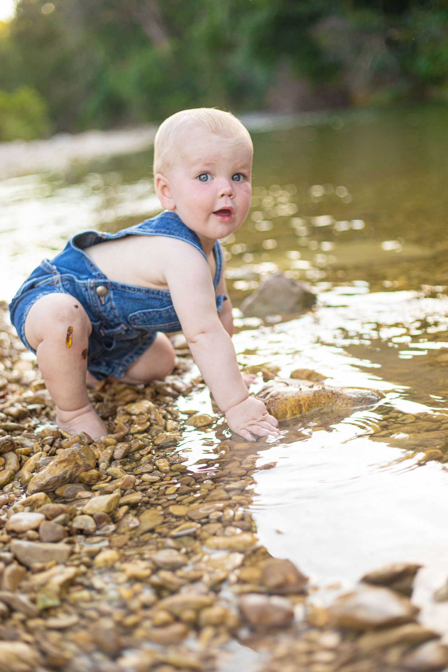 Baby in denim overalls crawling on wet river rocks.