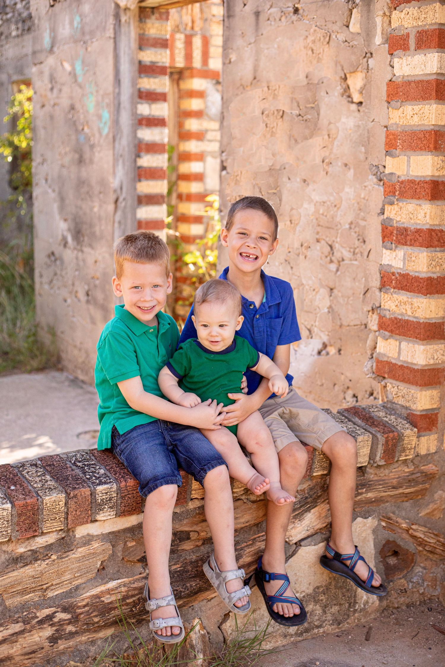 Three children smiling and laughing while sitting on brick ledge against rustic stone wall.