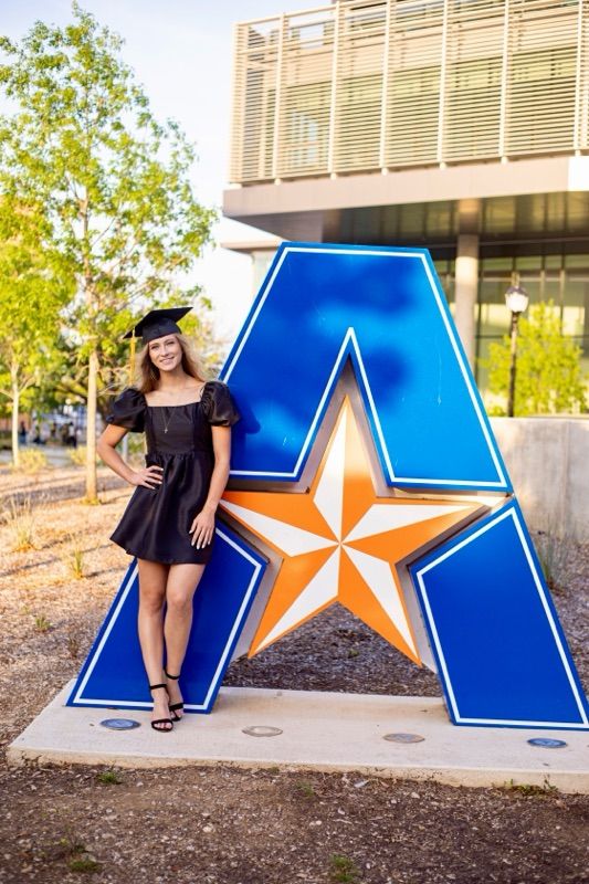 Graduate poses next to large blue UTA logo with orange star on campus.