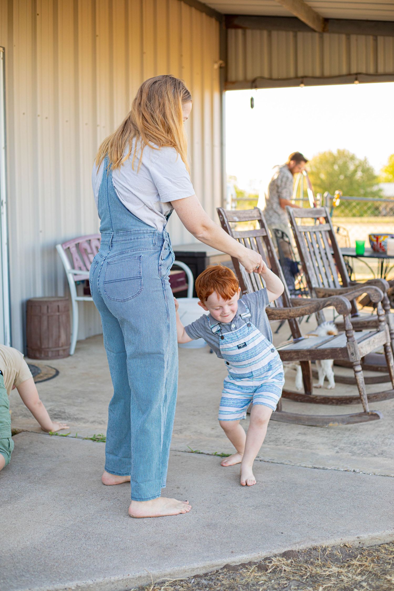 Woman in overalls playing with a red-haired toddler on a rustic farm porch with rocking chairs.