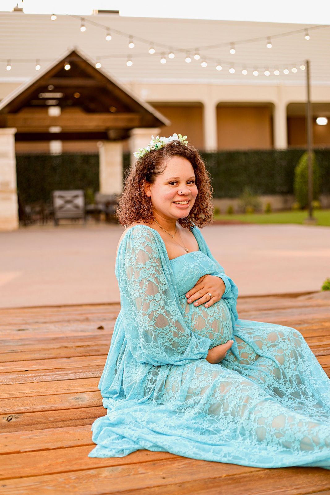 Pregnant woman in teal lace maternity gown with floral crown sits on wooden deck, smiling warmly at camera.