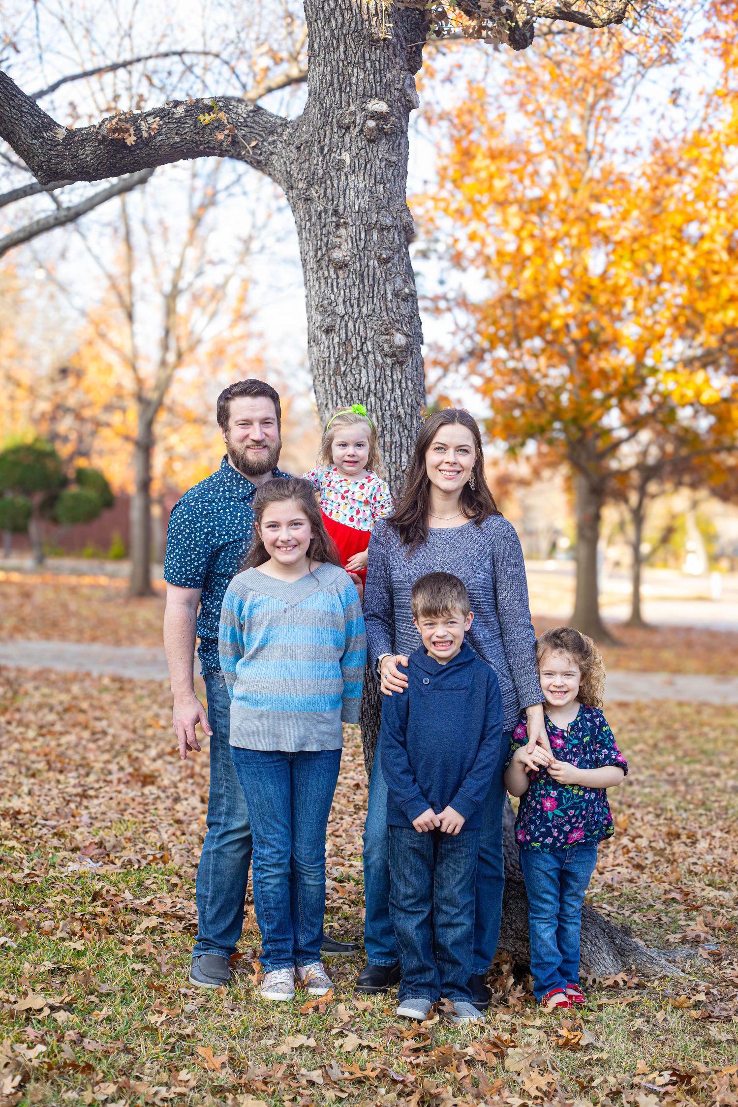 Family of six posing outdoors in autumn park with colorful fall foliage and large tree backdrop.