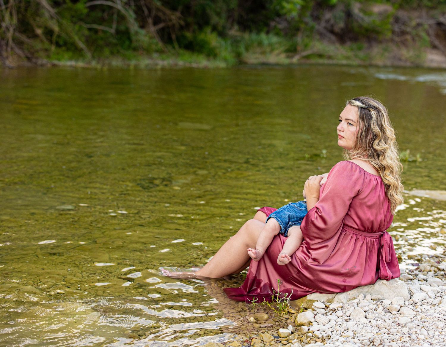 A peaceful riverside scene with someone in a pink dress sitting on the rocky shore.
