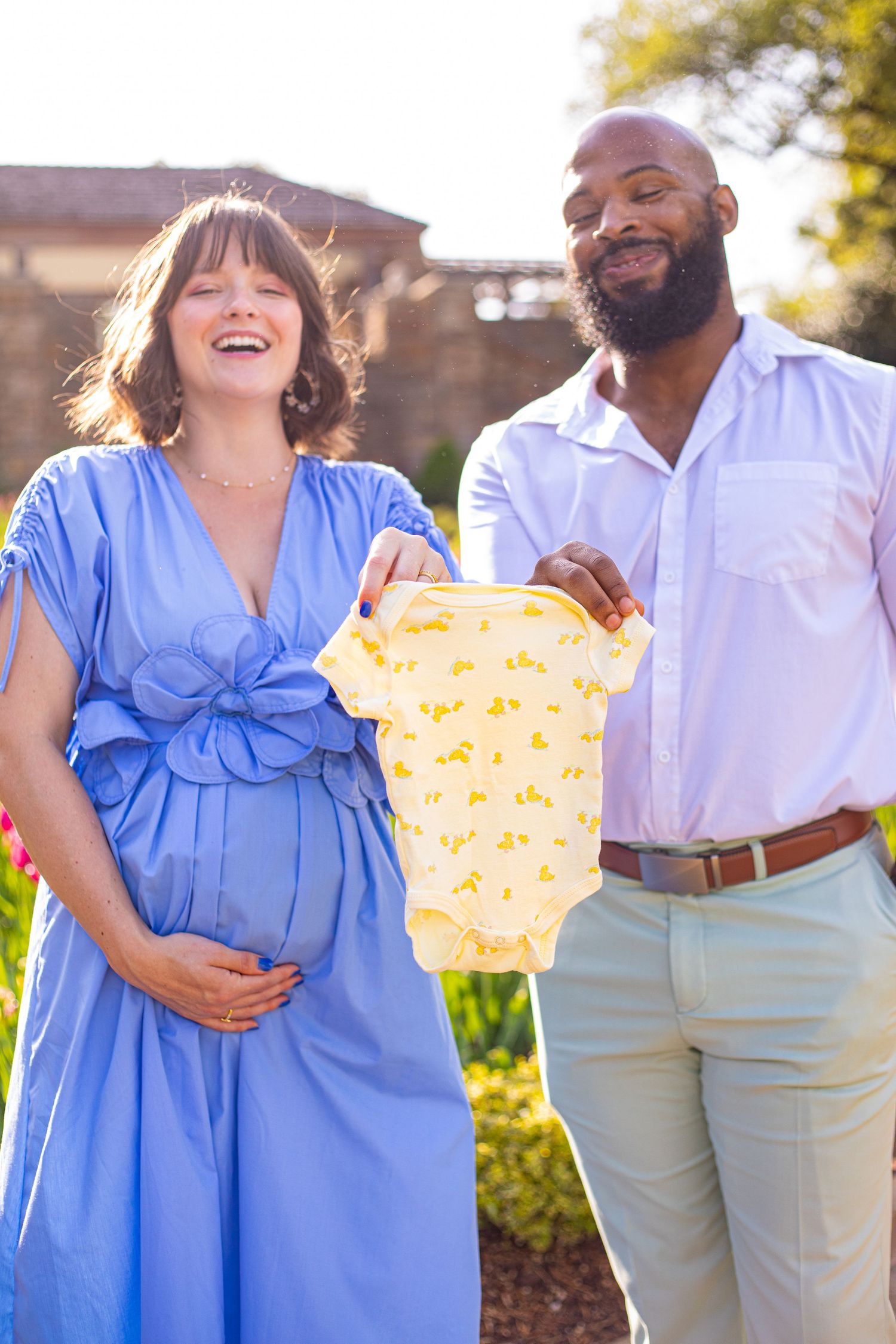 A couple holding up a yellow baby onesie in a sunny garden setting.