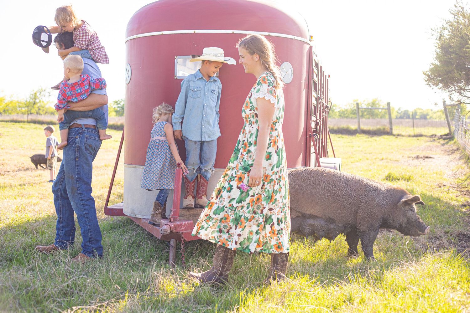 A group gathers around a large red barn structure in a grassy field at sunset.
