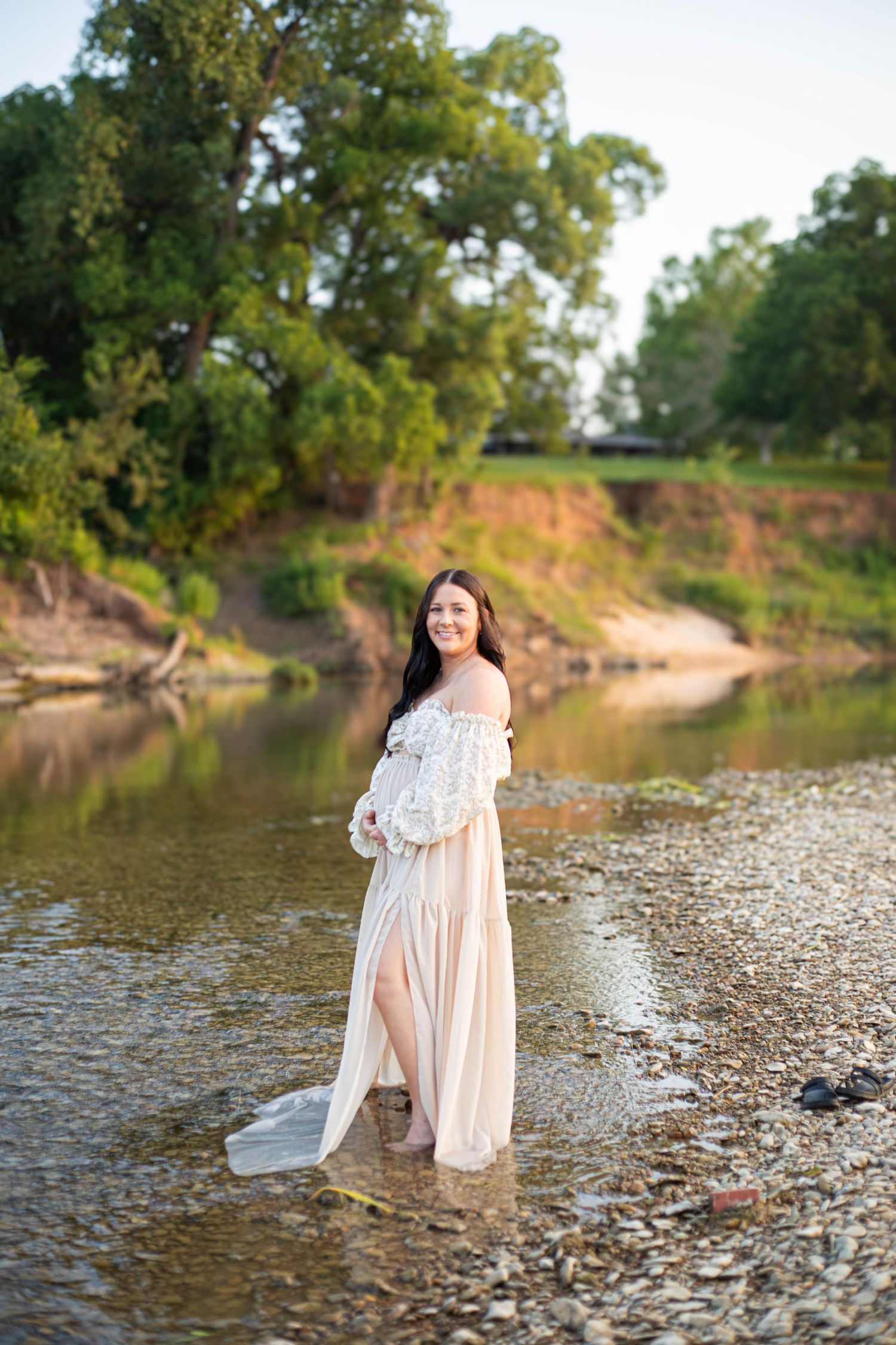 Pregnant woman in white lace and sheer gown standing in shallow river with green trees in background.