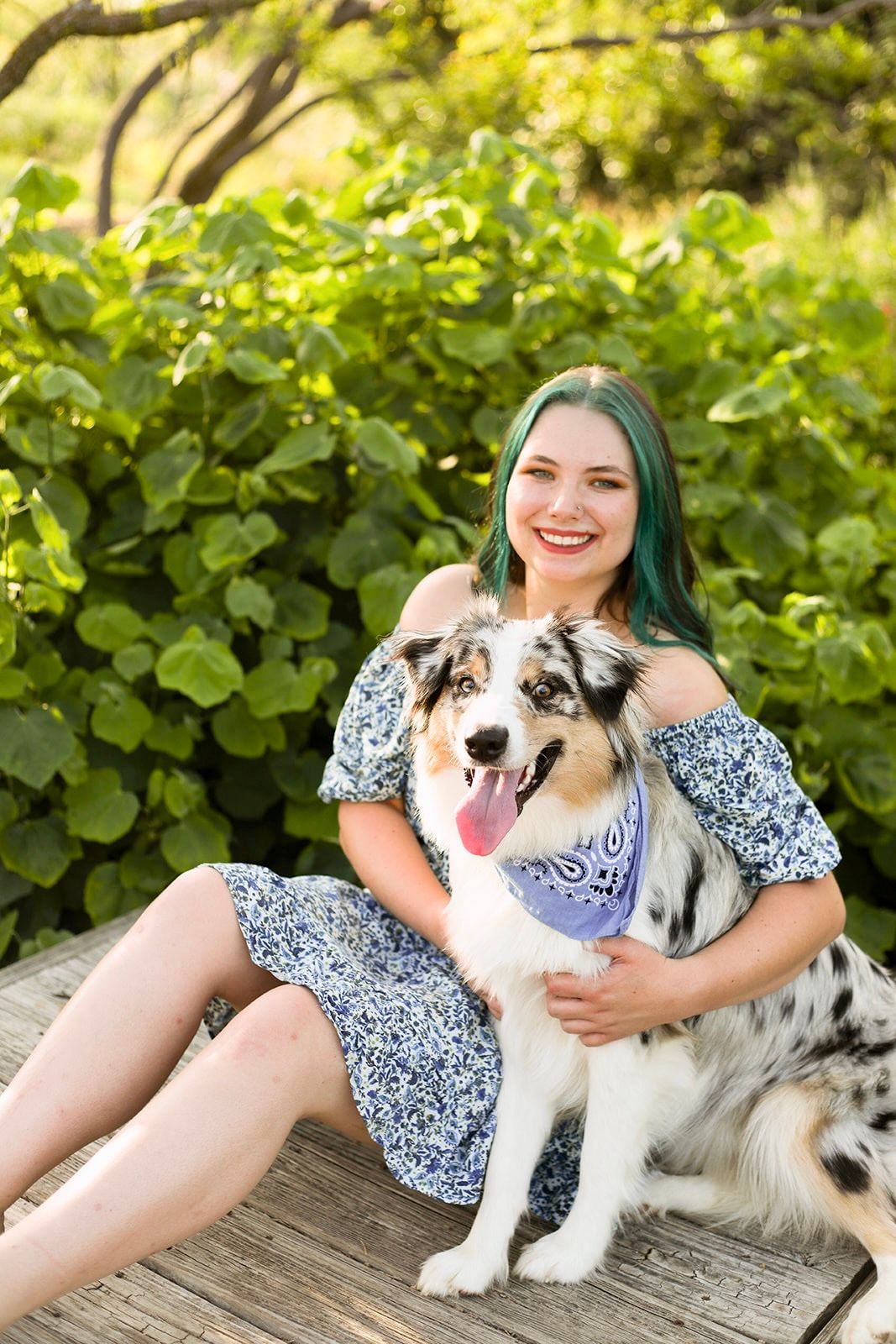 Person in floral dress sitting with Australian Shepherd dog on wooden bench outdoors.