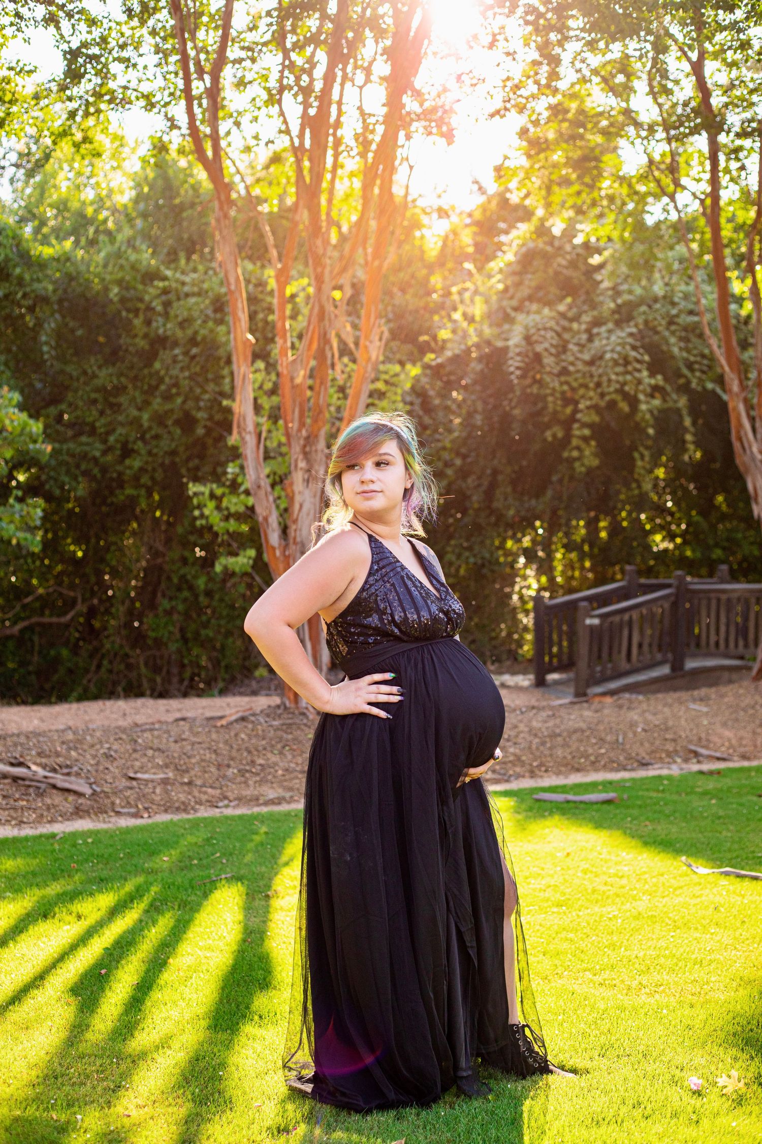 Pregnant woman in elegant black gown posing outdoors in sunlit garden with lush green trees.