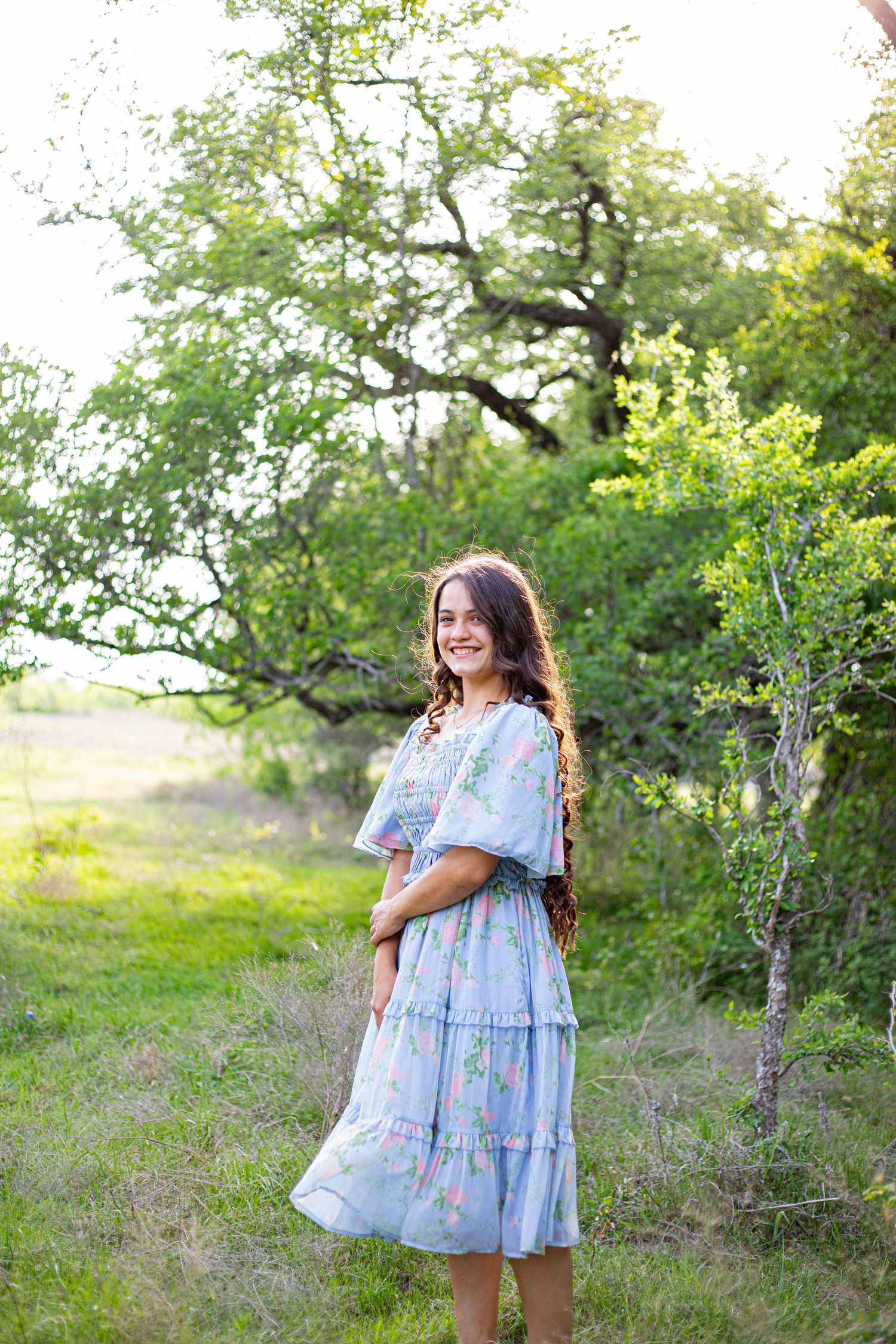 Smiling girl in light blue floral dress standing outdoors among lush green trees and grass.