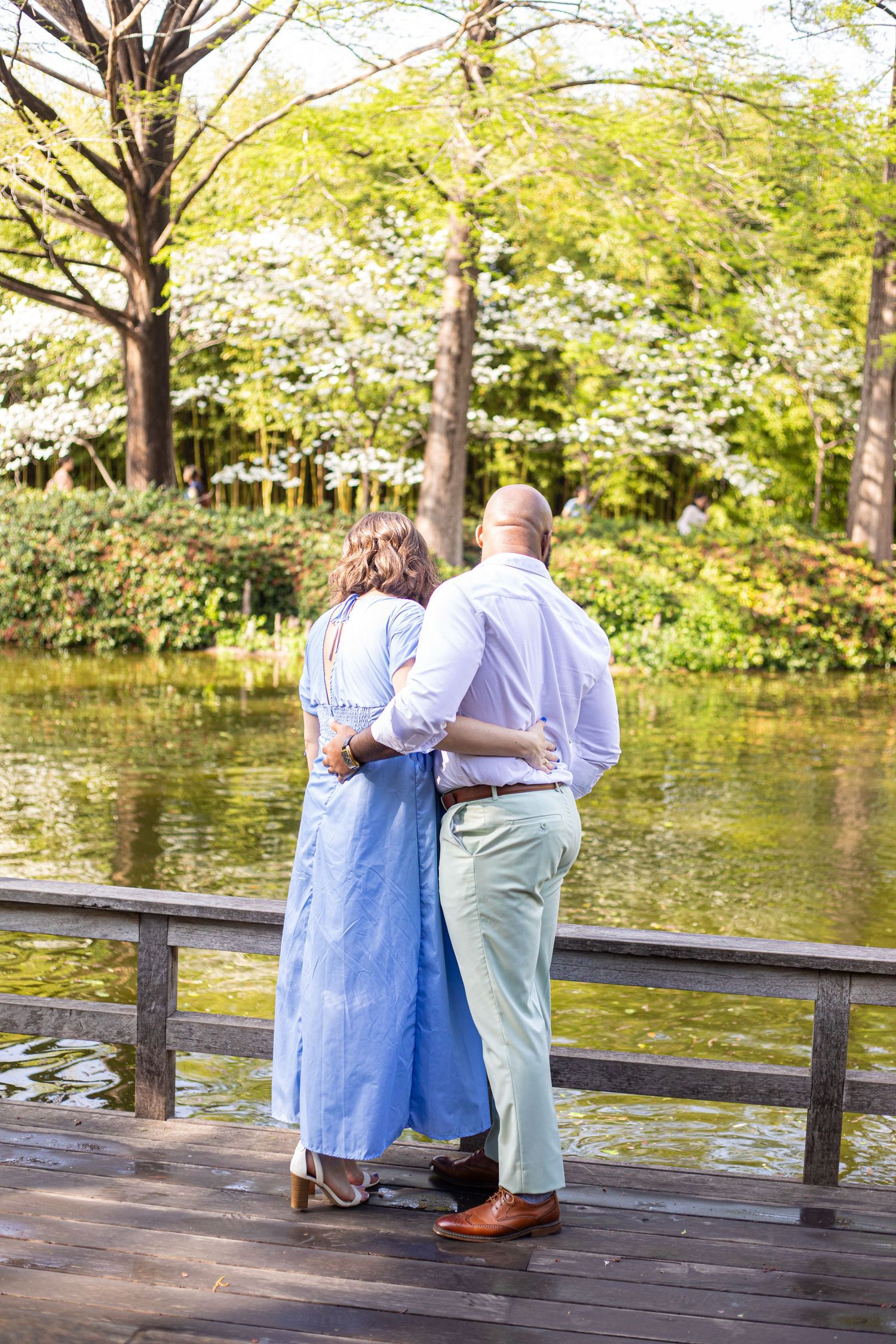 Couple walking together on wooden boardwalk by serene pond in park setting.