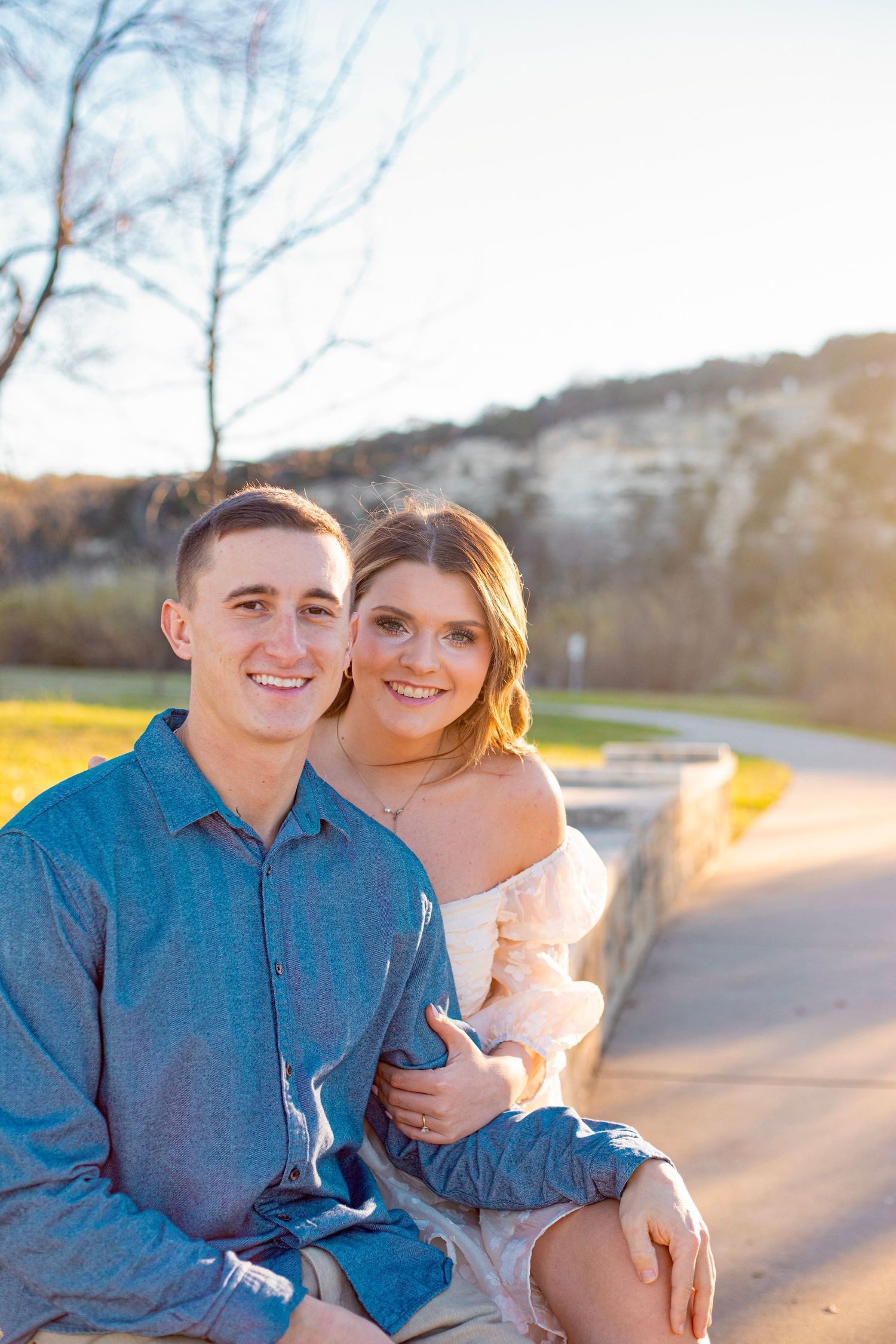 A couple sits together on a path during golden hour with scenic autumn landscape in background.