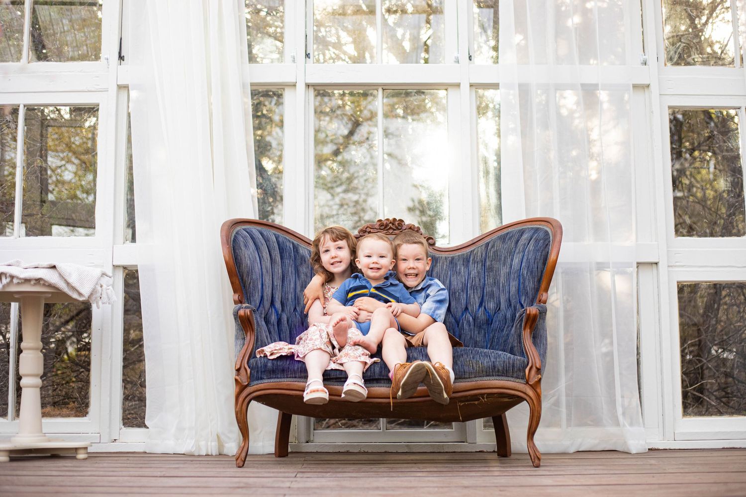 Three young children sitting together on a vintage blue velvet sofa in a bright sunlit room with white curtains.