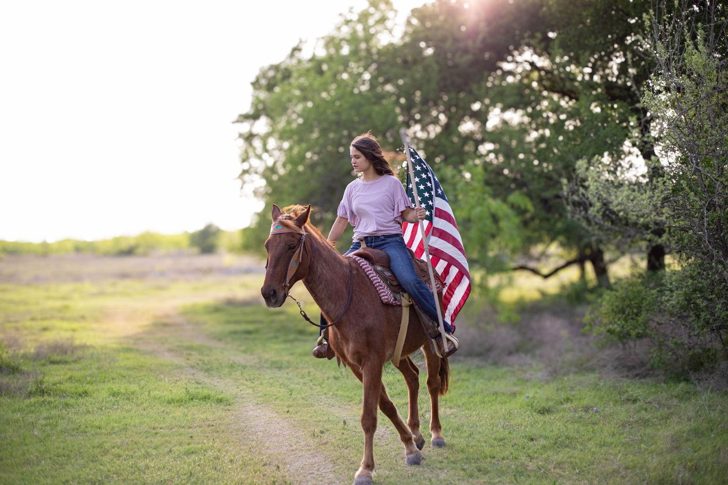 Woman riding a brown horse on a rural path while holding an American flag, surrounded by green trees and golden sunlight.