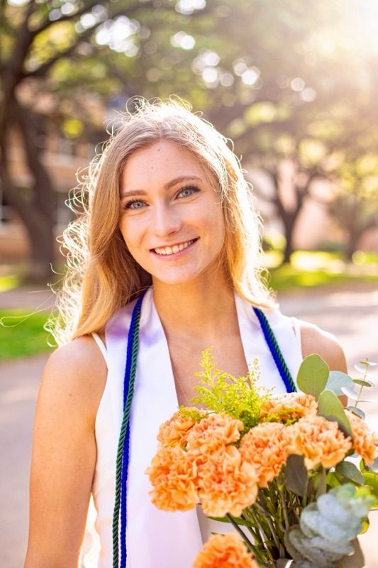 Graduate holding peach flowers with graduation stole in sunny outdoor setting.