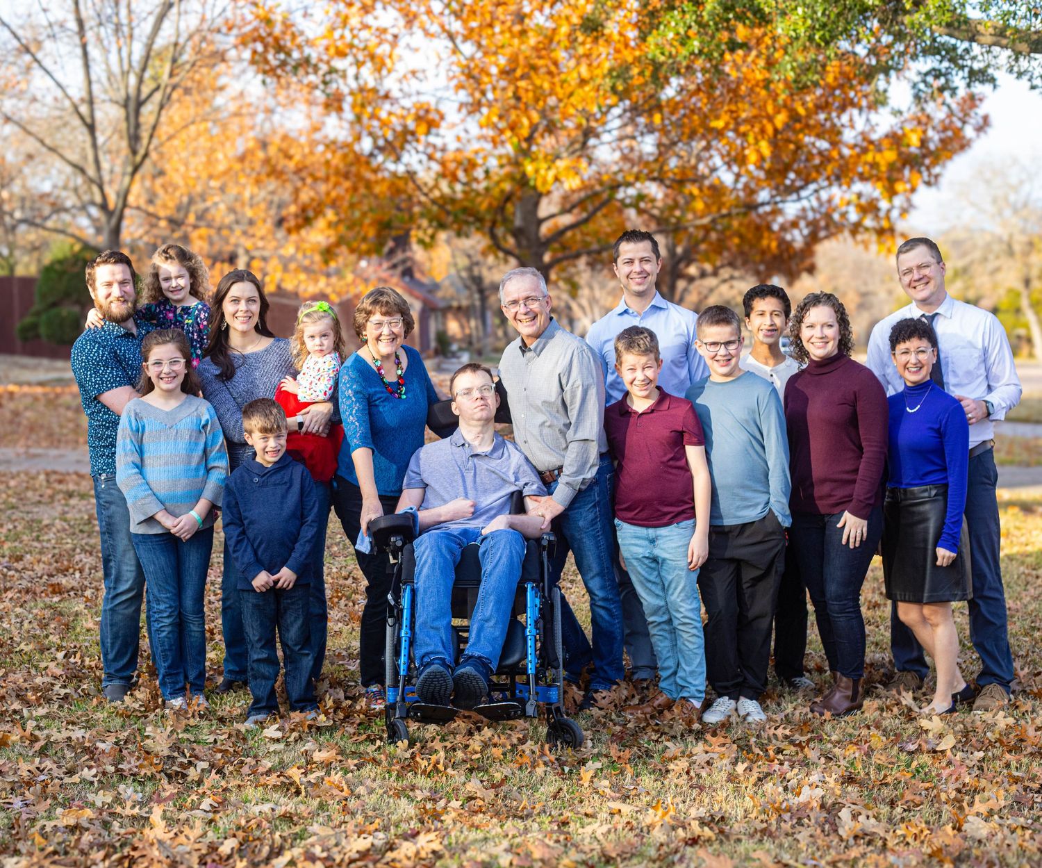 Large multigenerational family posing outdoors in autumn, with colorful fall foliage and a child in a wheelchair at center.