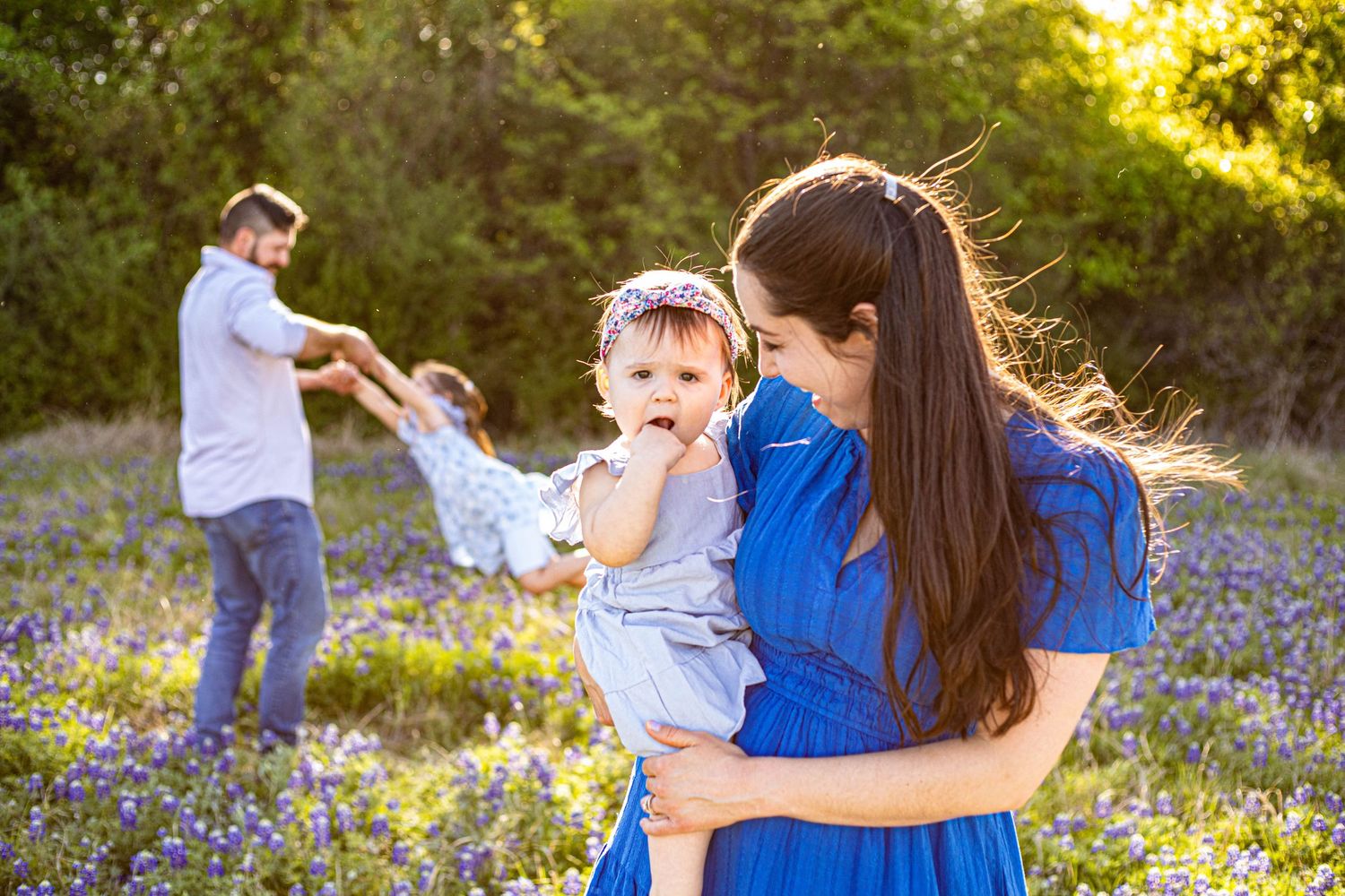 A mother in a blue dress holds her baby in a wildflower field while a father swings their child in the background.