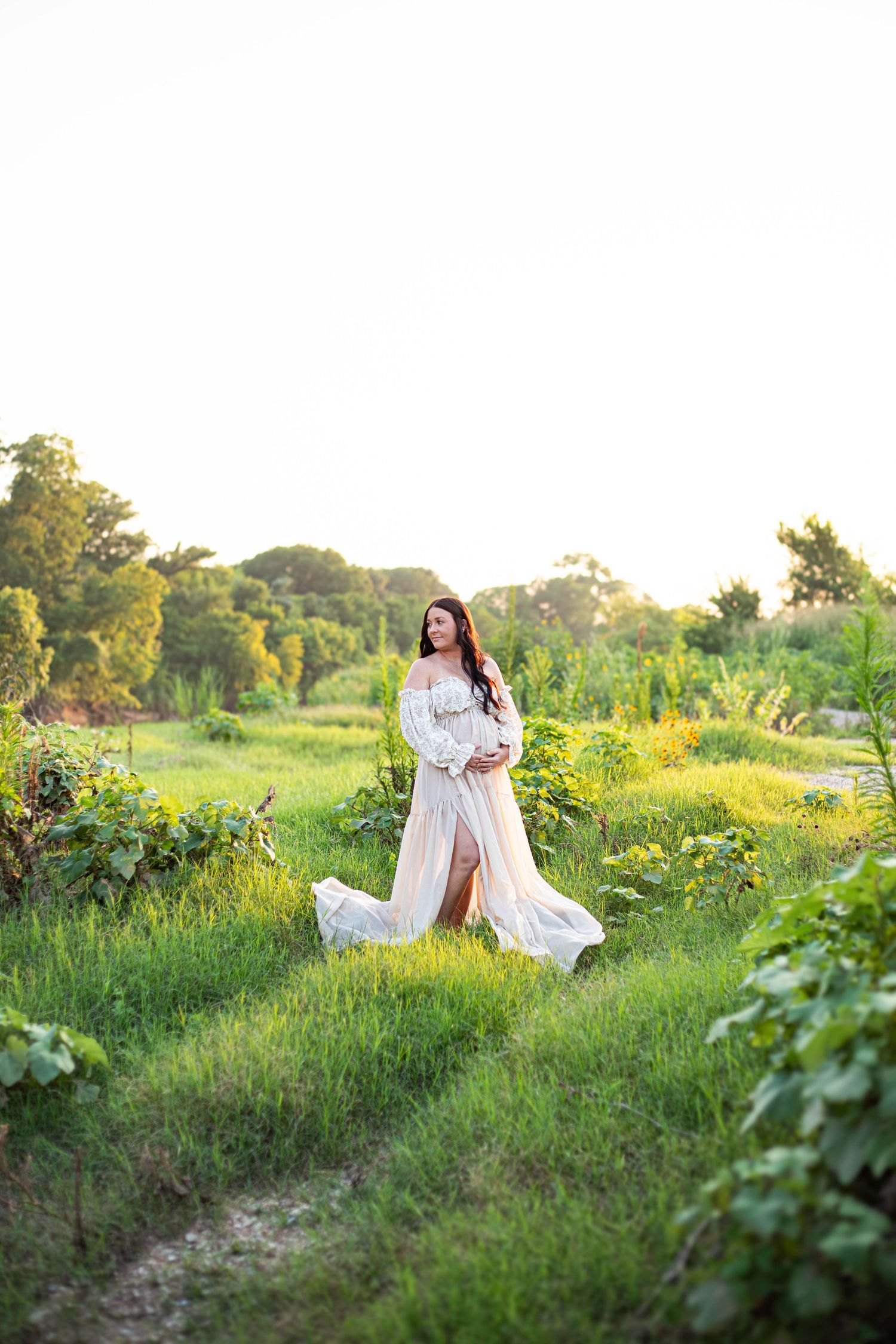 Pregnant woman in flowing white lace gown standing in lush green field during golden hour sunset.