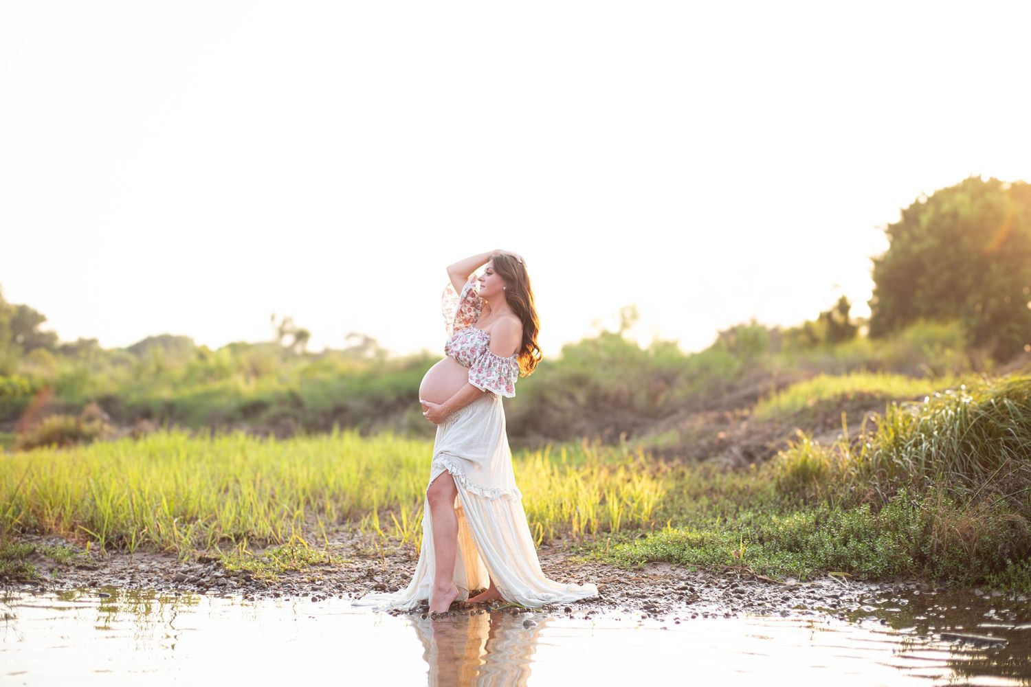 Pregnant woman in white flowy gown standing in shallow water surrounded by golden sunlit greenery.