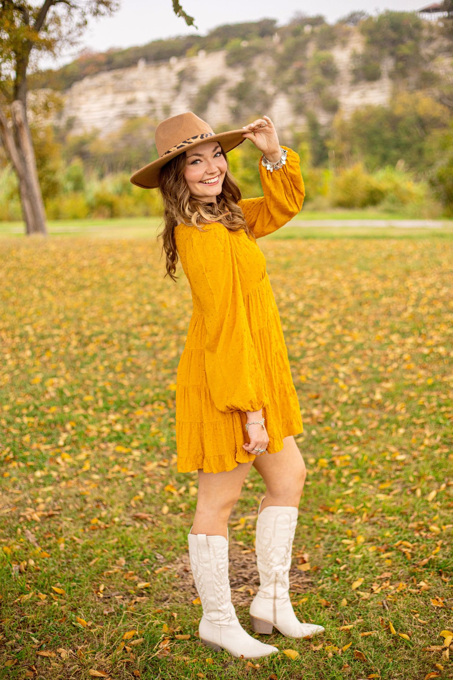 A person wearing a bright yellow dress and white boots poses in a field of yellow wildflowers.