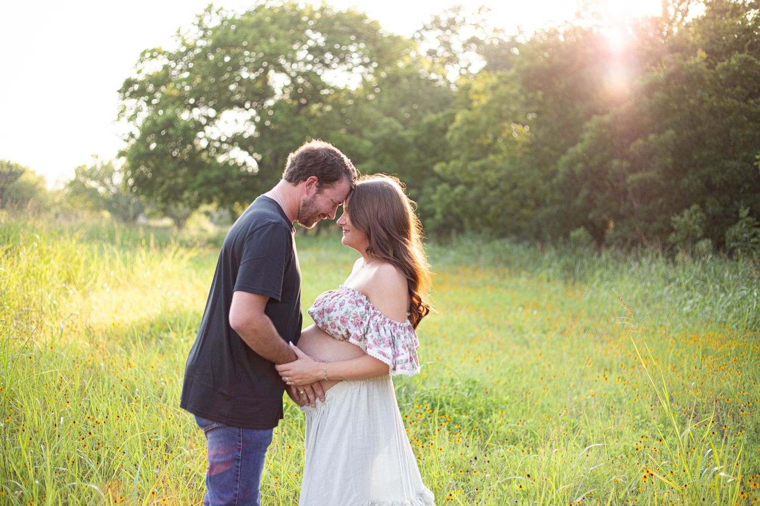 Couple sharing a romantic moment in a sunlit meadow, surrounded by lush green grass and trees.