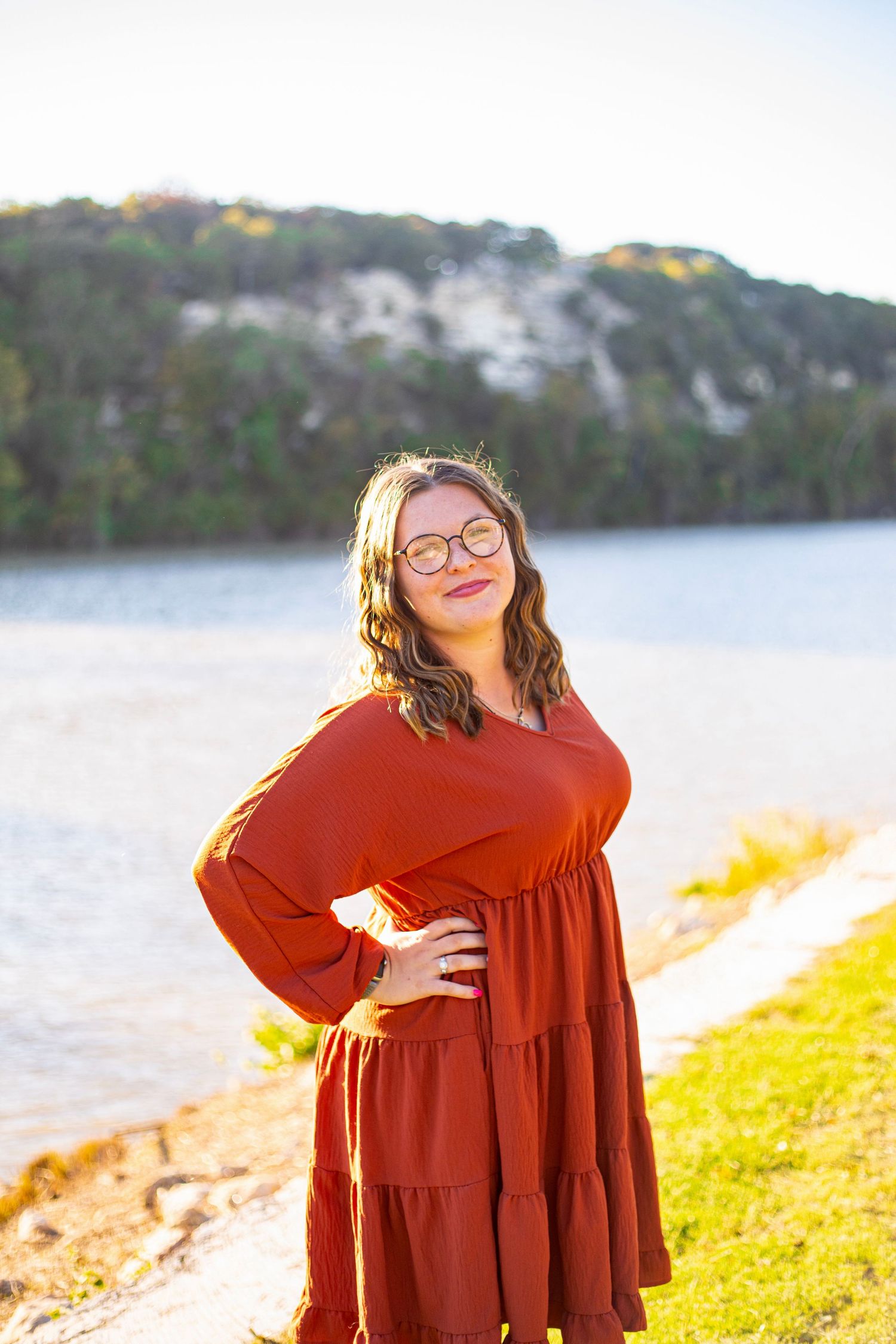 A person in a flowing red dress poses by a lakeside during golden hour.