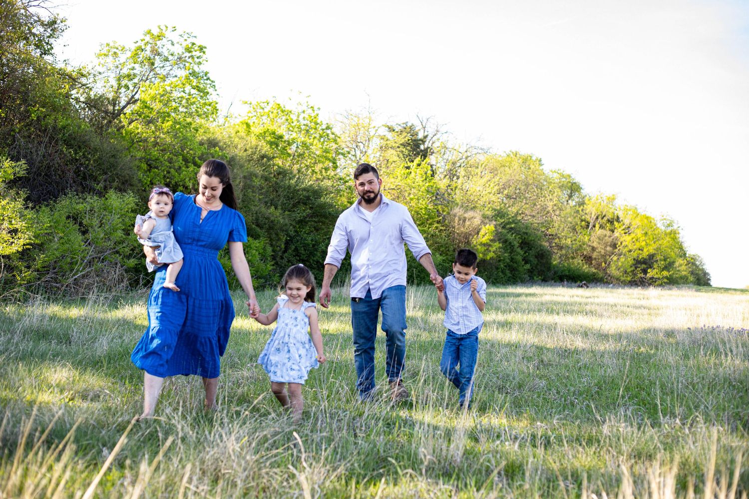 Family of five walking hand-in-hand through a sunny green meadow, parents with three young children in a nature setting.