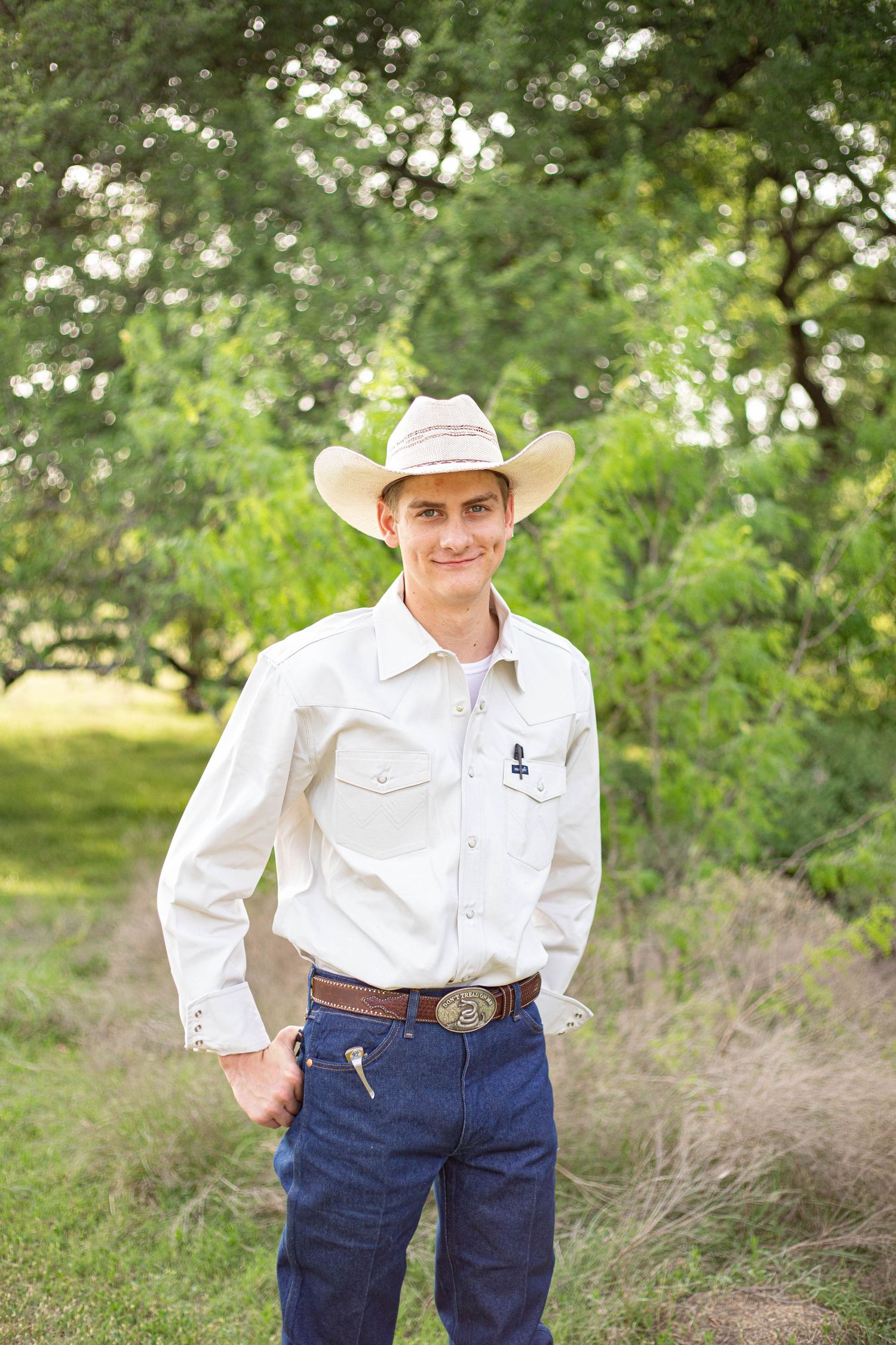 Young man in white western shirt, cowboy hat, and jeans smiling outdoors in a green tree-lined setting.