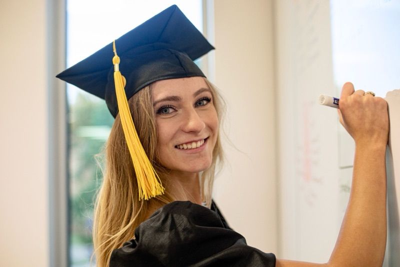 Graduate in academic regalia with yellow tassel against bright interior background.