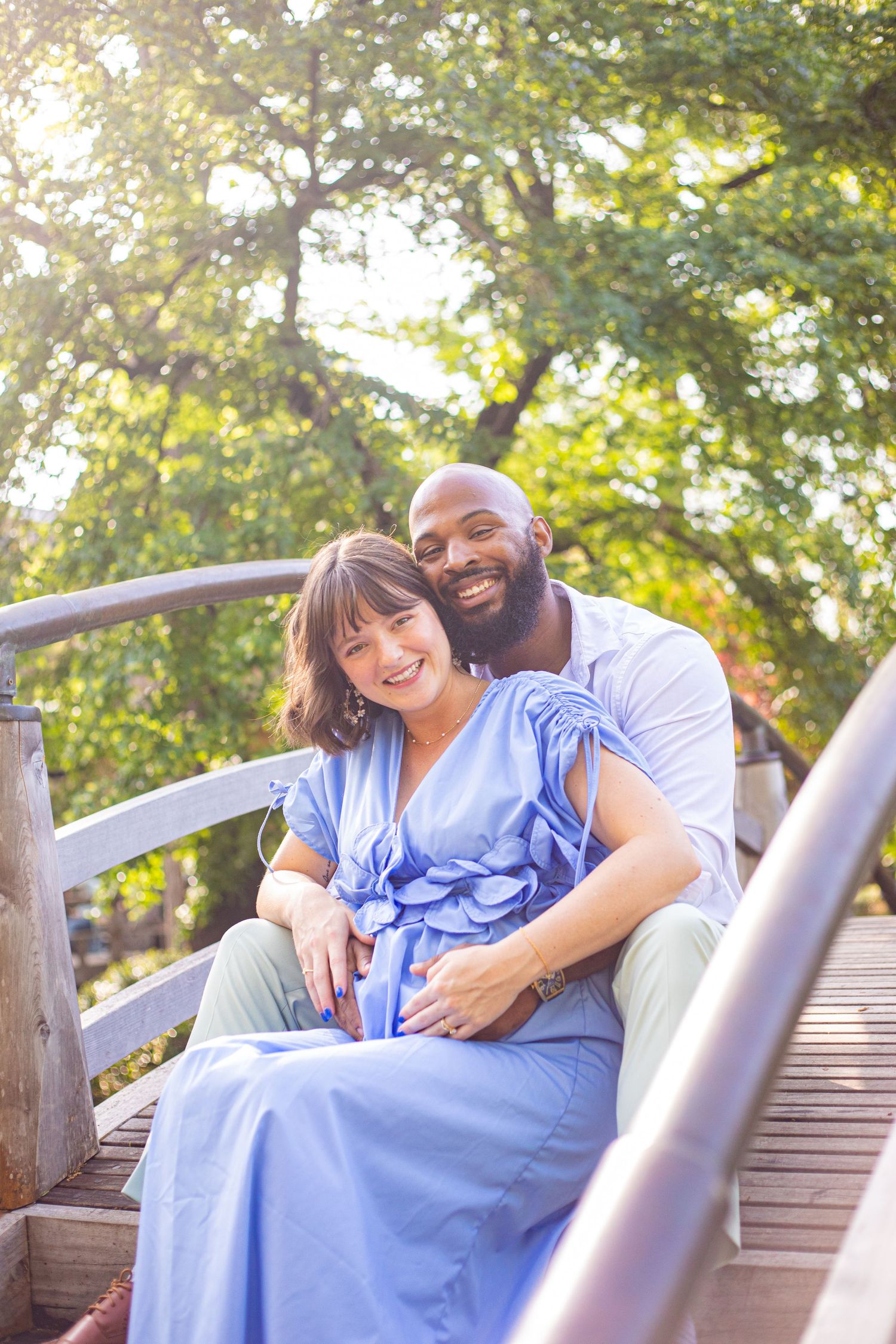 Happy couple lounging on wooden bridge railing in sunny garden setting.
