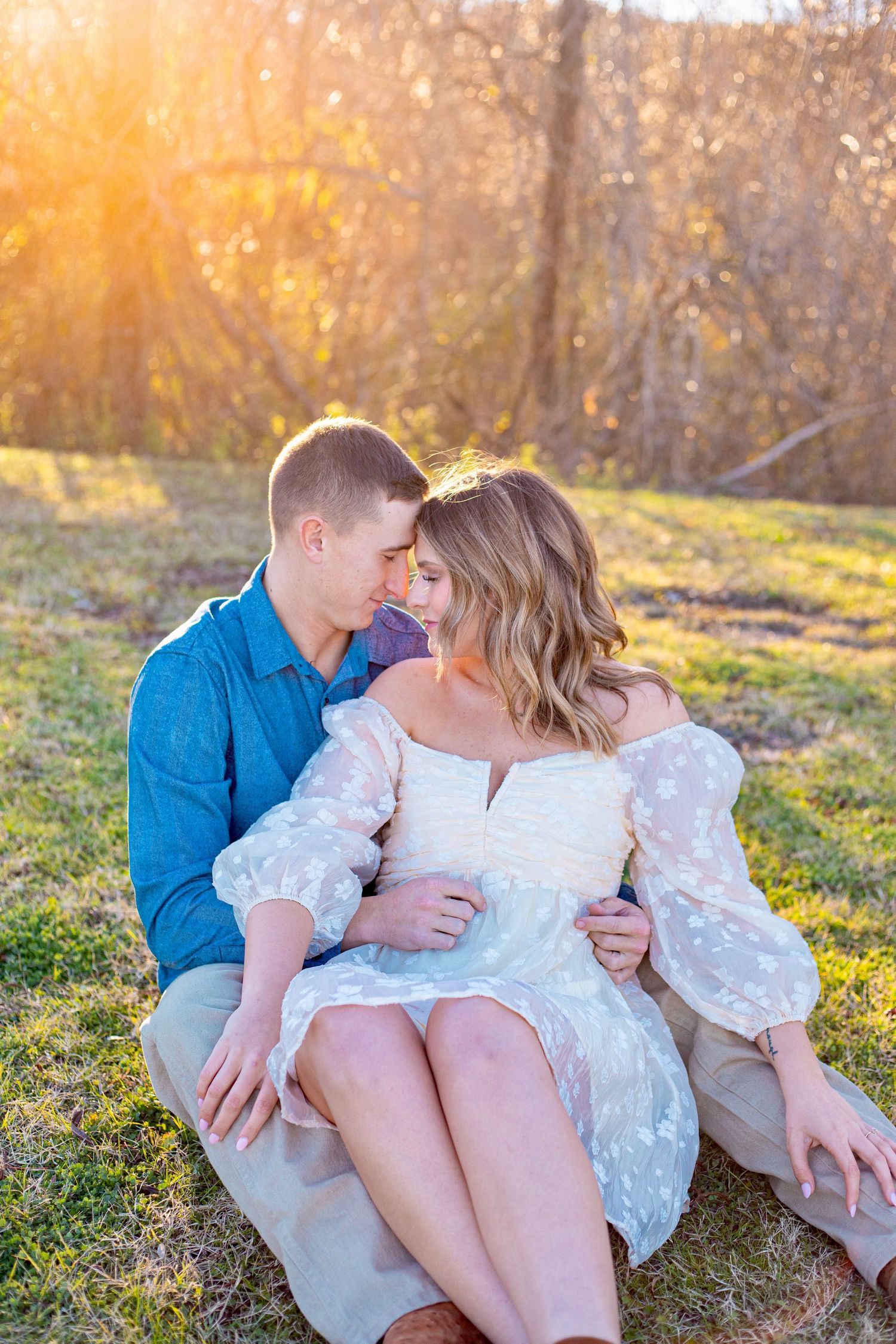 A romantic couple embraces while sitting on grass during sunset with golden light filtering through trees.