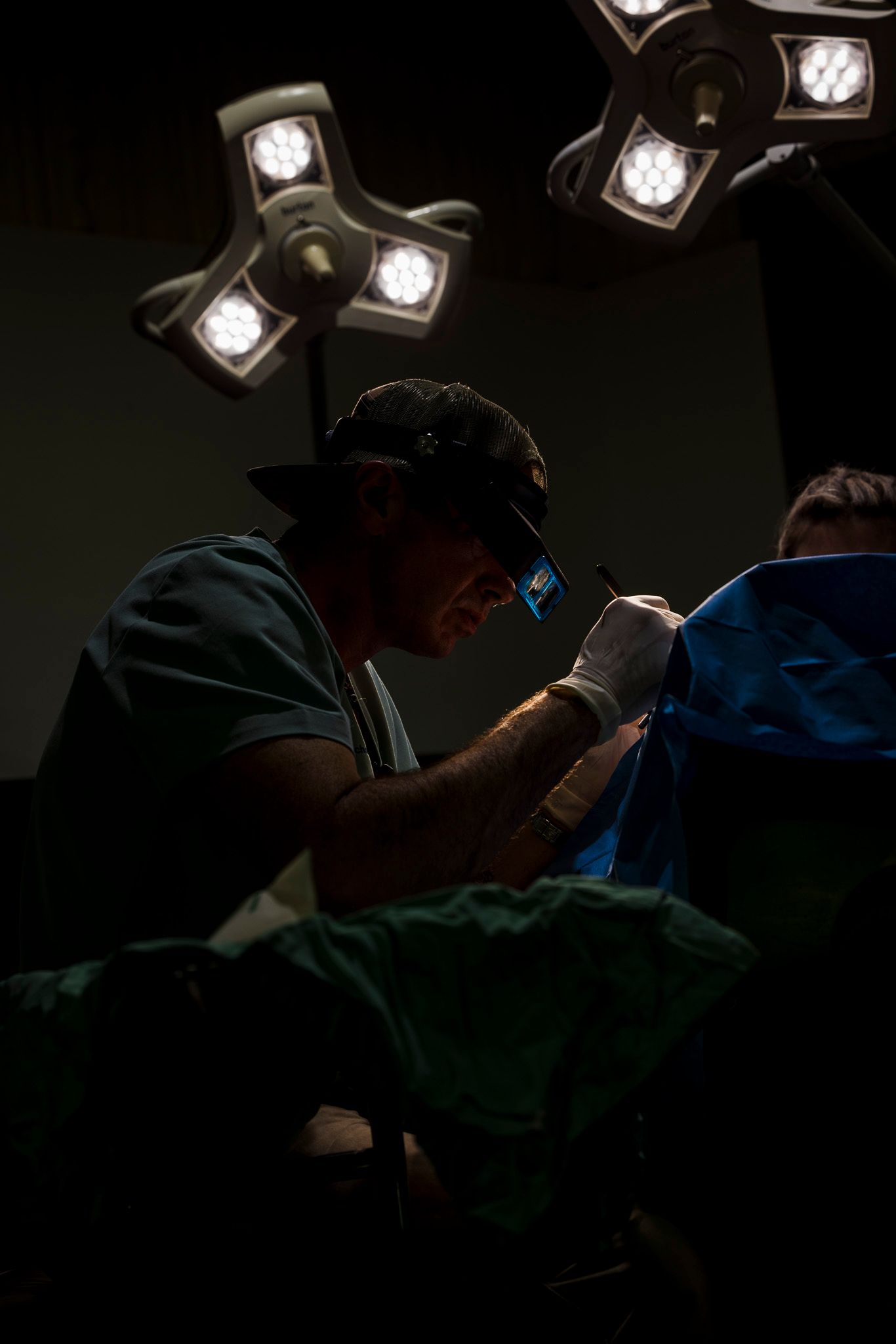 Veterinarian performing surgery in operating room under bright surgical lights in dark dramatic lighting.