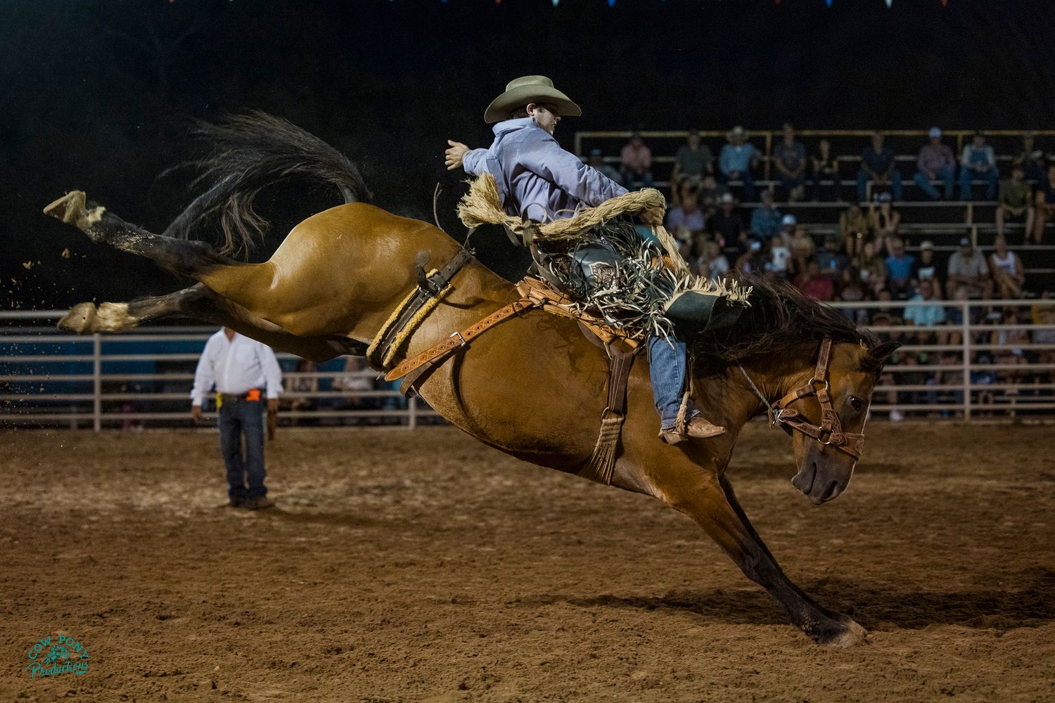 A rodeo rider in western attire competes in bronc riding competition on a bucking horse in a dirt arena.