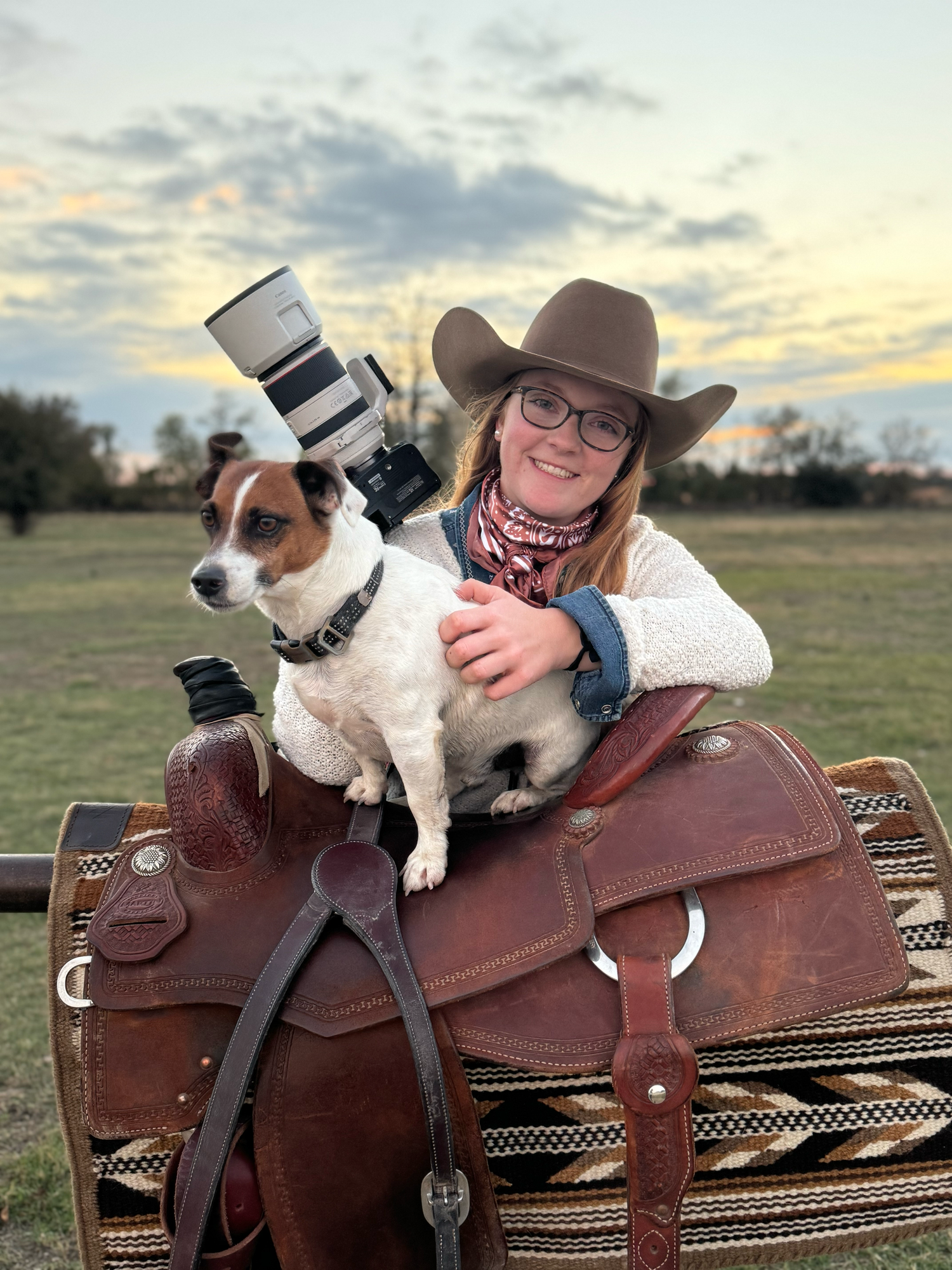 A Jack Russell Terrier sits on a western saddle with a camera lens while someone in cowgirl attire poses at sunset.