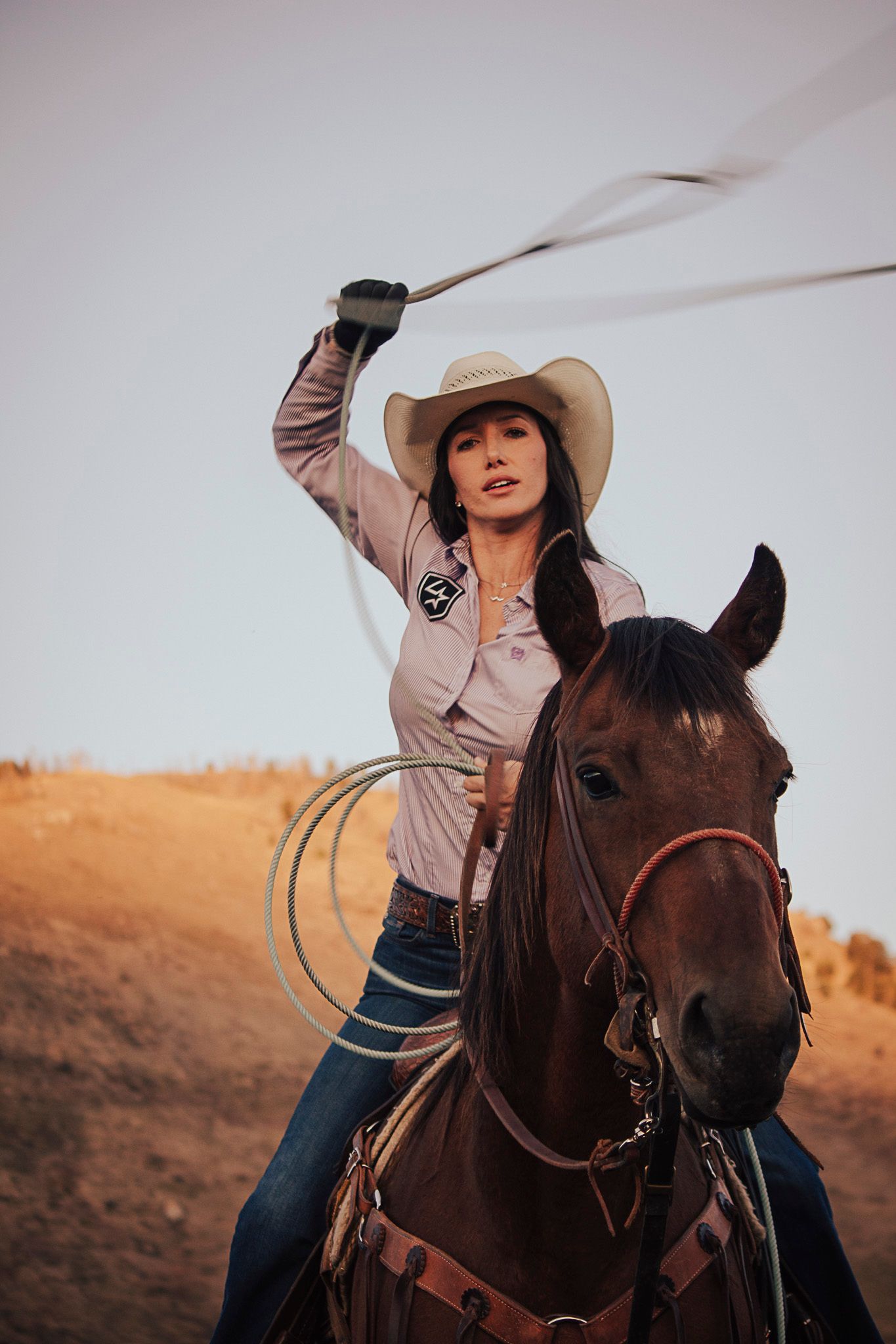 A rider in a cowboy hat practicing with a rope while mounted on a bay horse against a mountain landscape at sunset in Jackson Hole, Wyoming