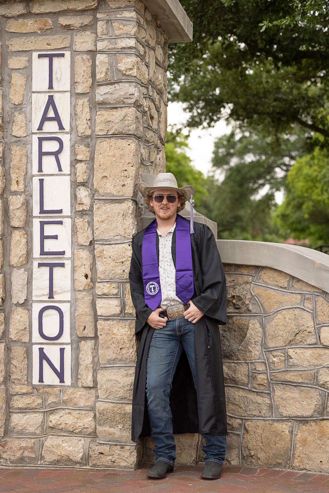 Graduate in black graduation gown with purple stole stands by stone Tarleton University entrance pillar.