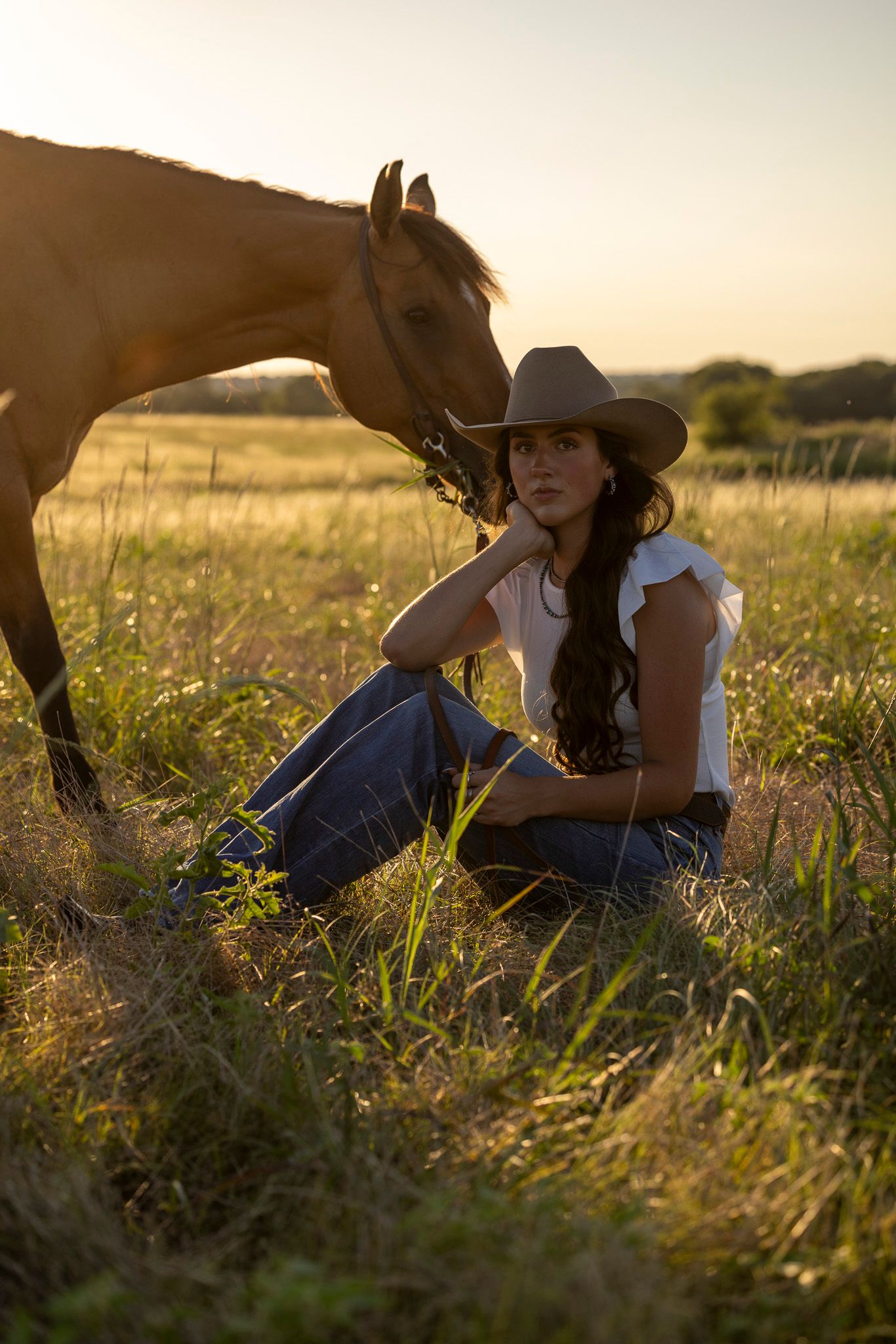 Senior portraits of a horse and rider rest in a golden field at sunset, surrounded by tall grass and warm evening light.