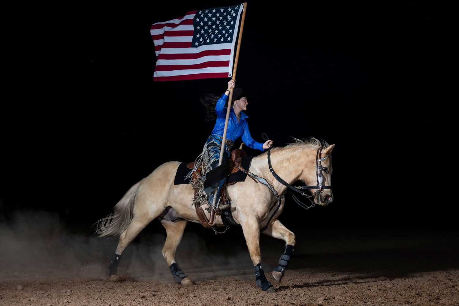 A horse rider in blue carries the American flag while galloping on a palomino horse at night.