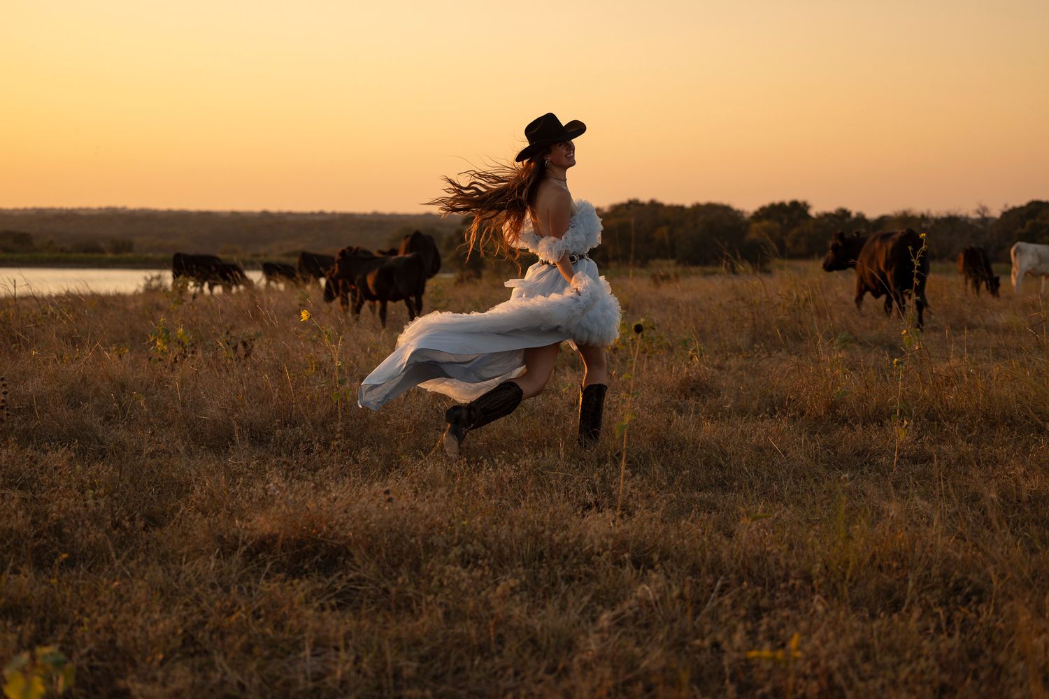 Person in flowing pale blue dress frolics gracefully in meadow at sunset with cattle grazing in background.