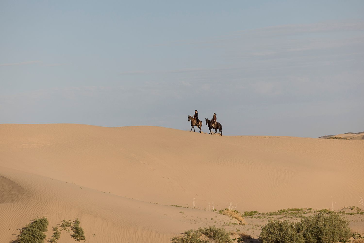 Two people riding horses across golden sand dunes in a vast desert landscape. Little Sahara Sand Dunes in Utah.