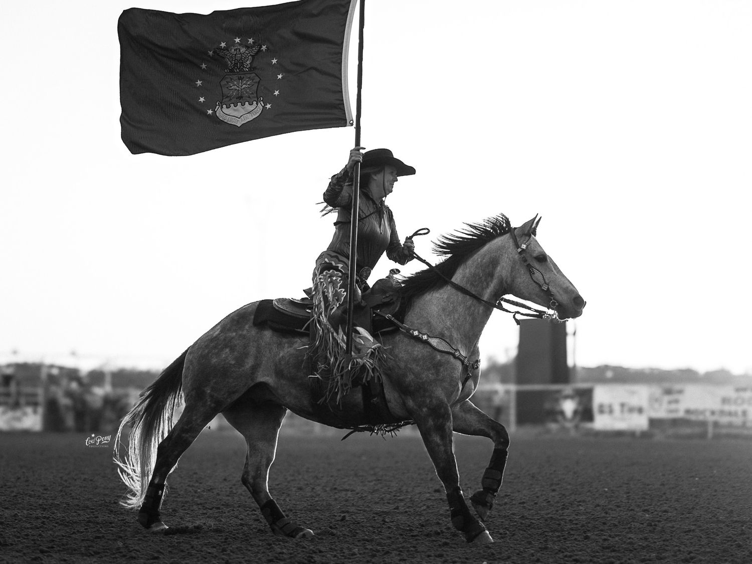 A black and white photo shows a rider on horseback carrying a US Air Force flag across a rodeo arena during opening ceremonies.