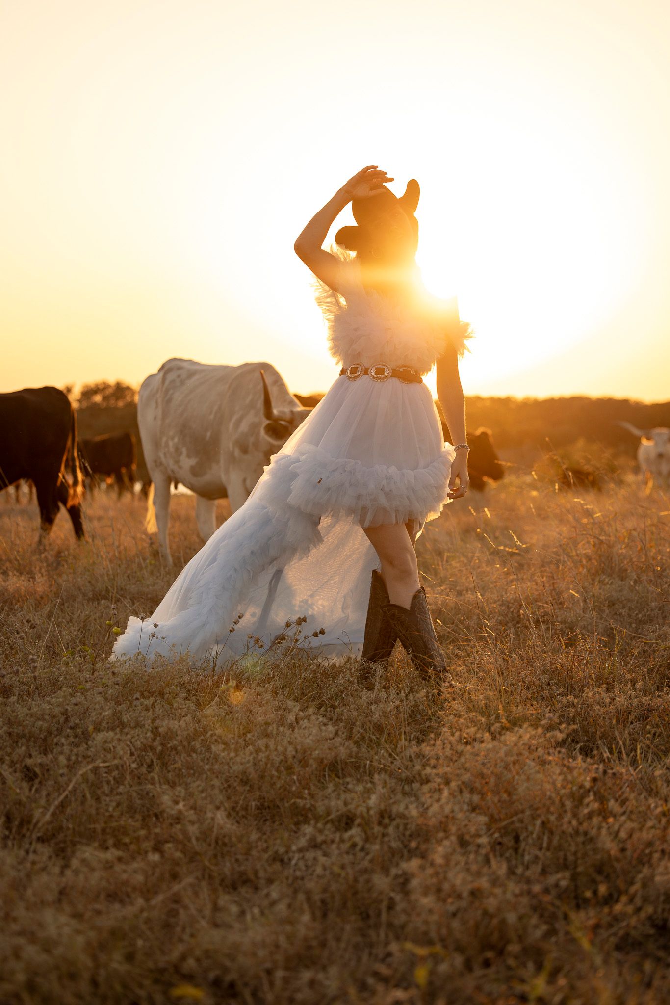 Woman in flowing dress dancing in golden sunlit field with cattle grazing in background at sunset.